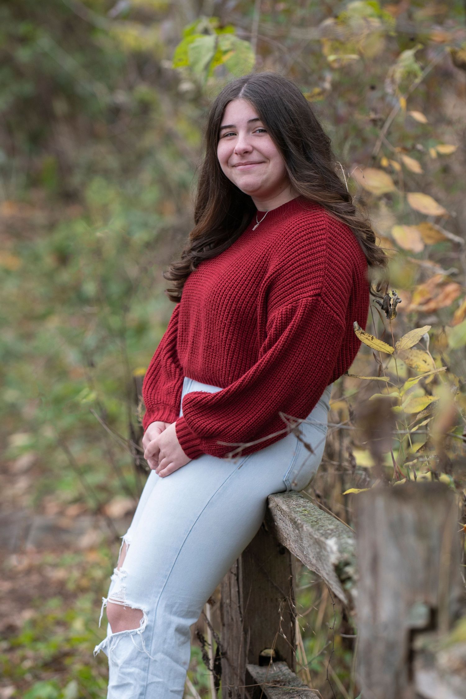Person in red sweater and light jeans poses outdoors against natural autumn foliage backdrop.