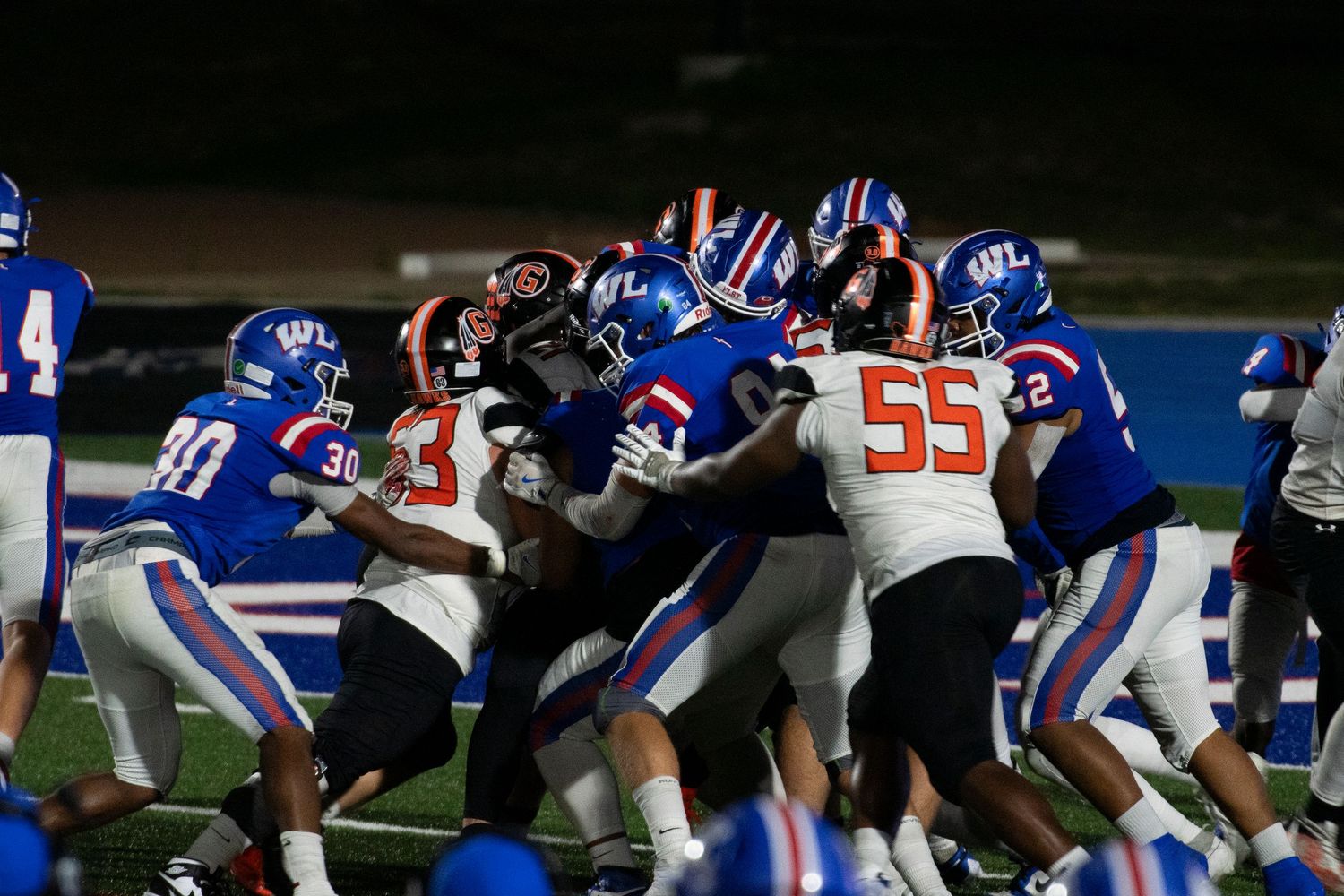 High school football players in blue and white uniforms clash against opponents in orange during an intense night game.