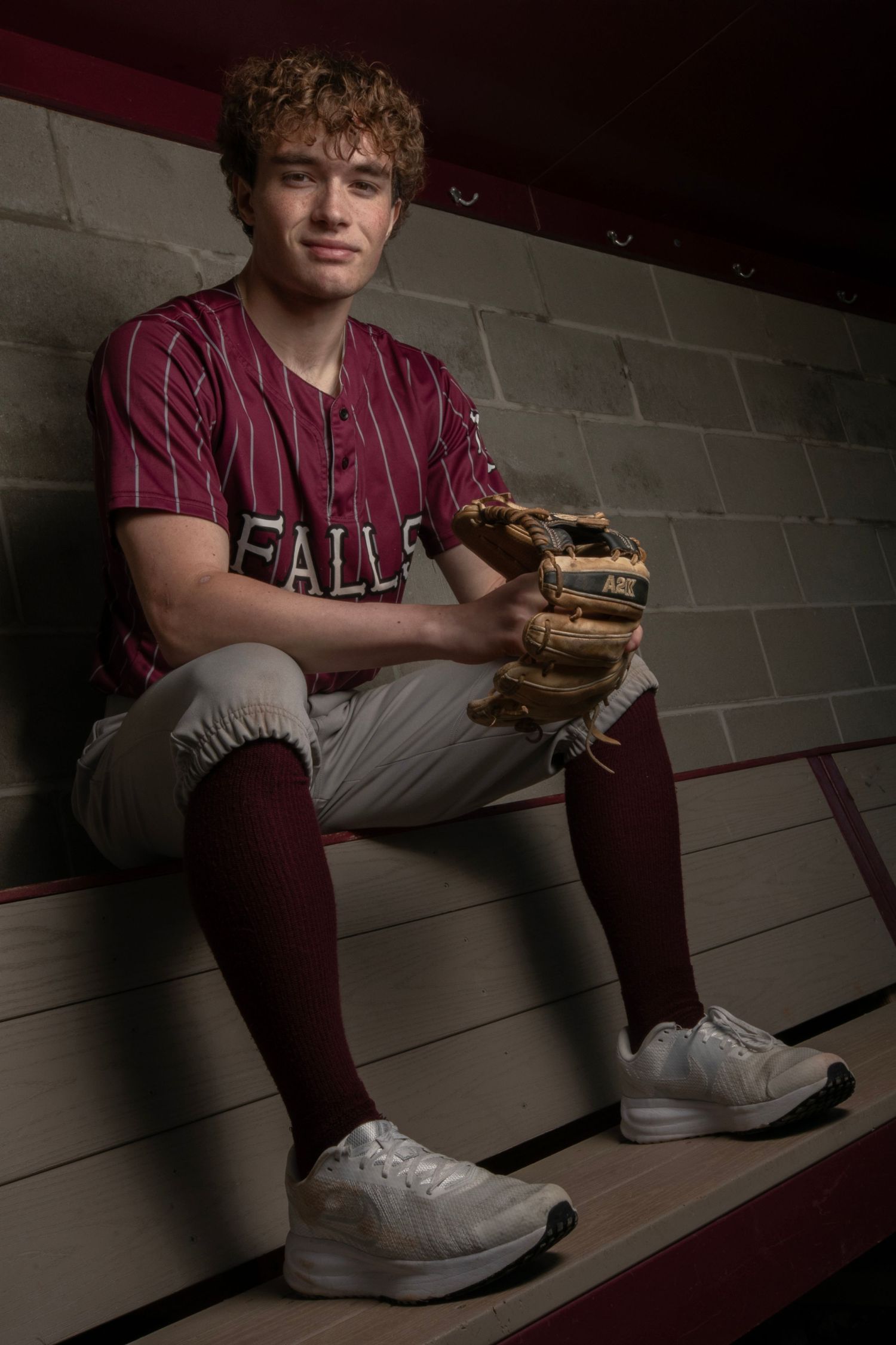Young baseball player in maroon pinstripe uniform sits in dugout holding a glove, wearing high socks and white cleats.