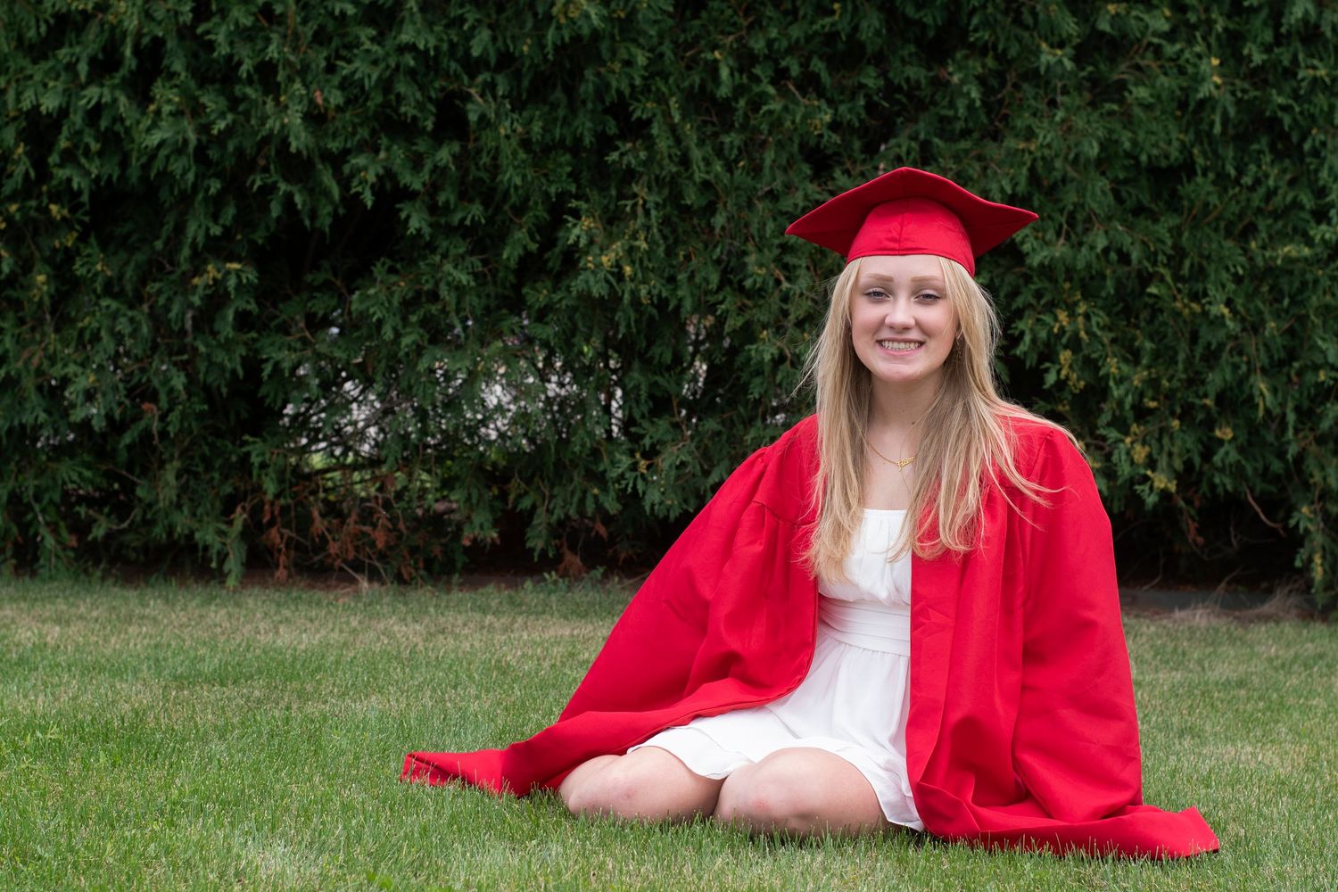 A person wearing a red graduation cap and gown sits outdoors against a backdrop of green foliage.