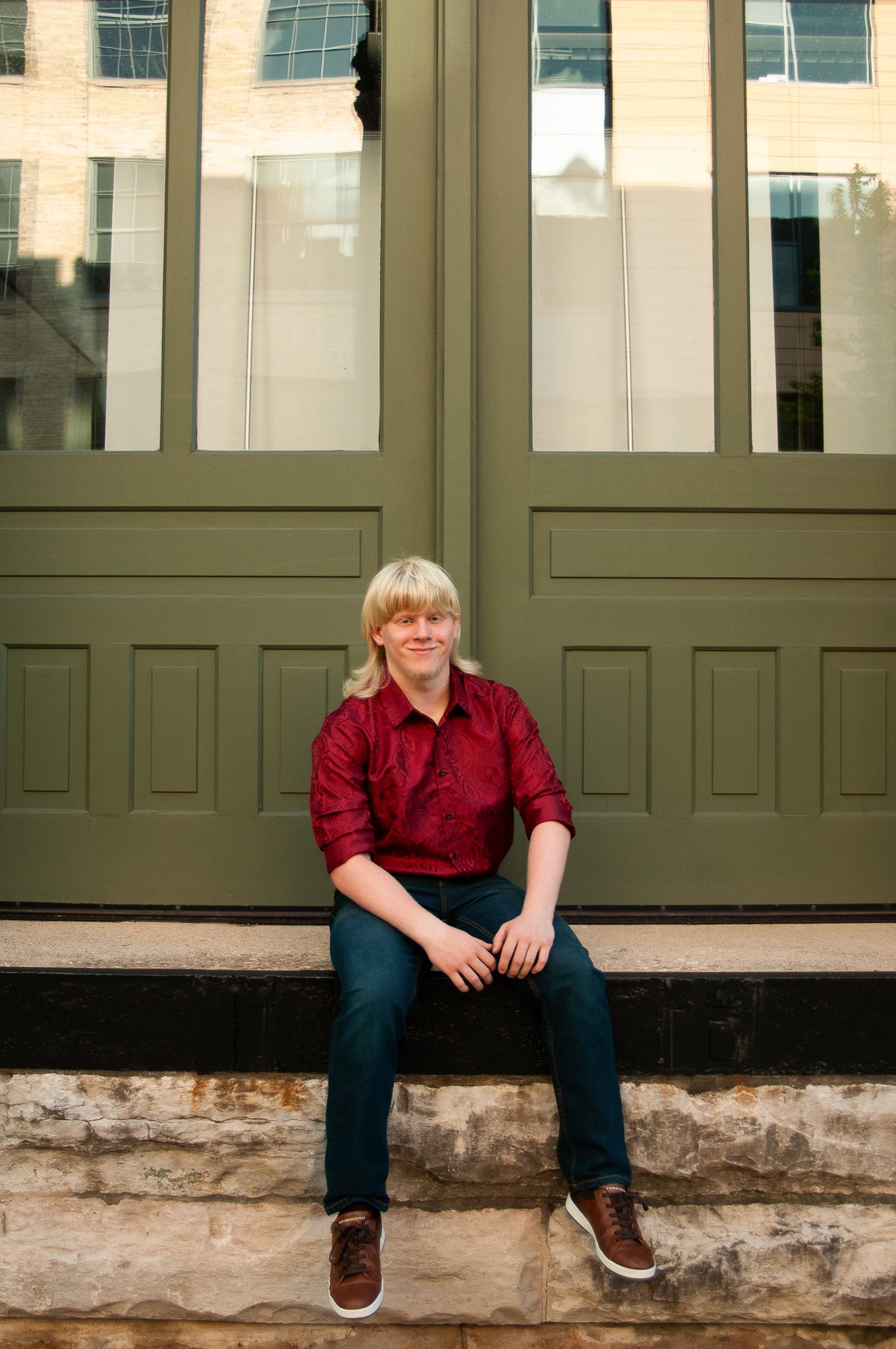 High school senior poses casually concrete ledge against a green paneled door in the Brewery District in Milwaukee, Wisconsin.