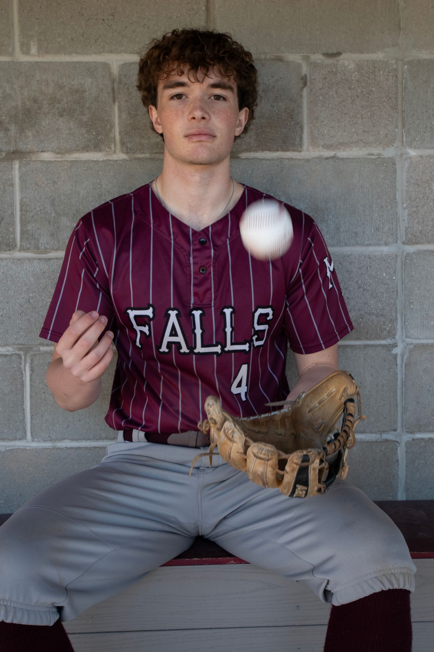 Young baseball player in maroon Falls jersey tossing a ball while seated in dugout wearing a glove.