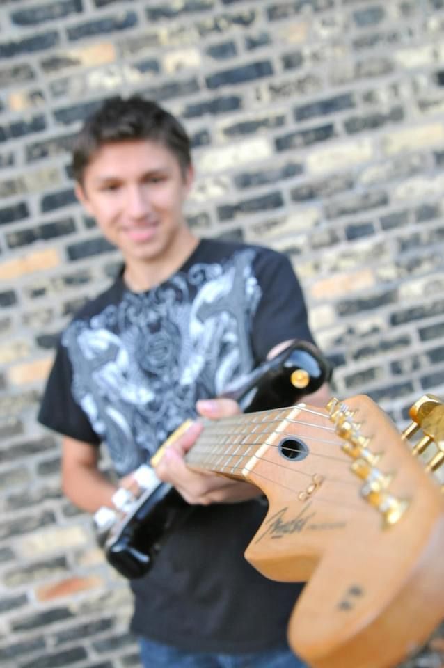 Young man smiling while holding a Fender electric guitar with gold tuners against a brick wall backdrop.
