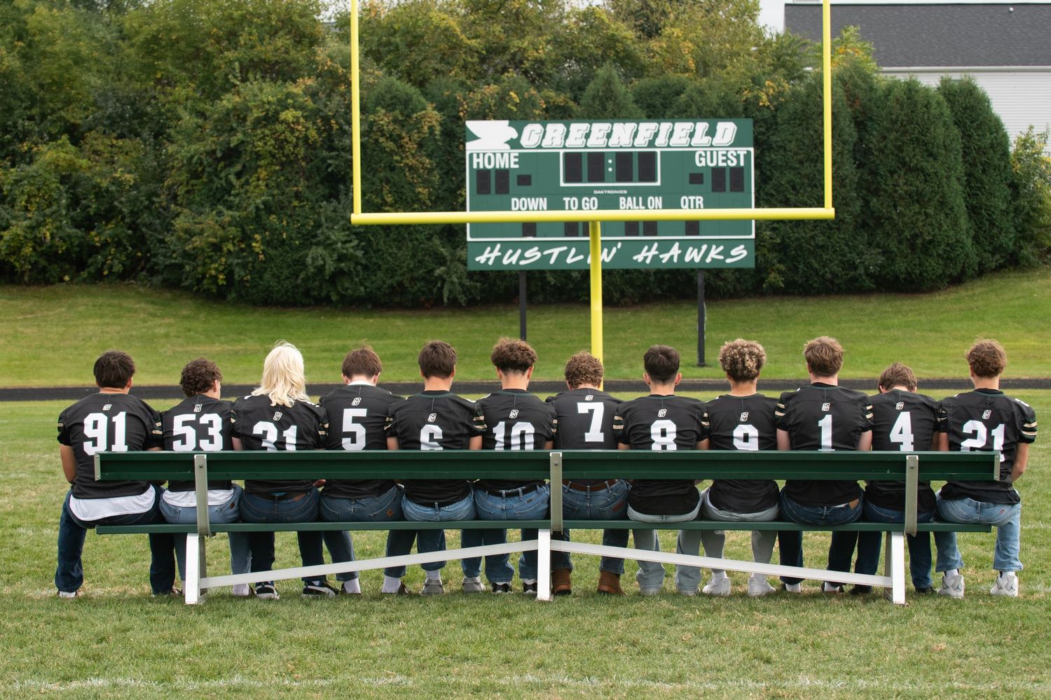 Football players sit on a bench watching a game on the field with goal posts visible in the background.