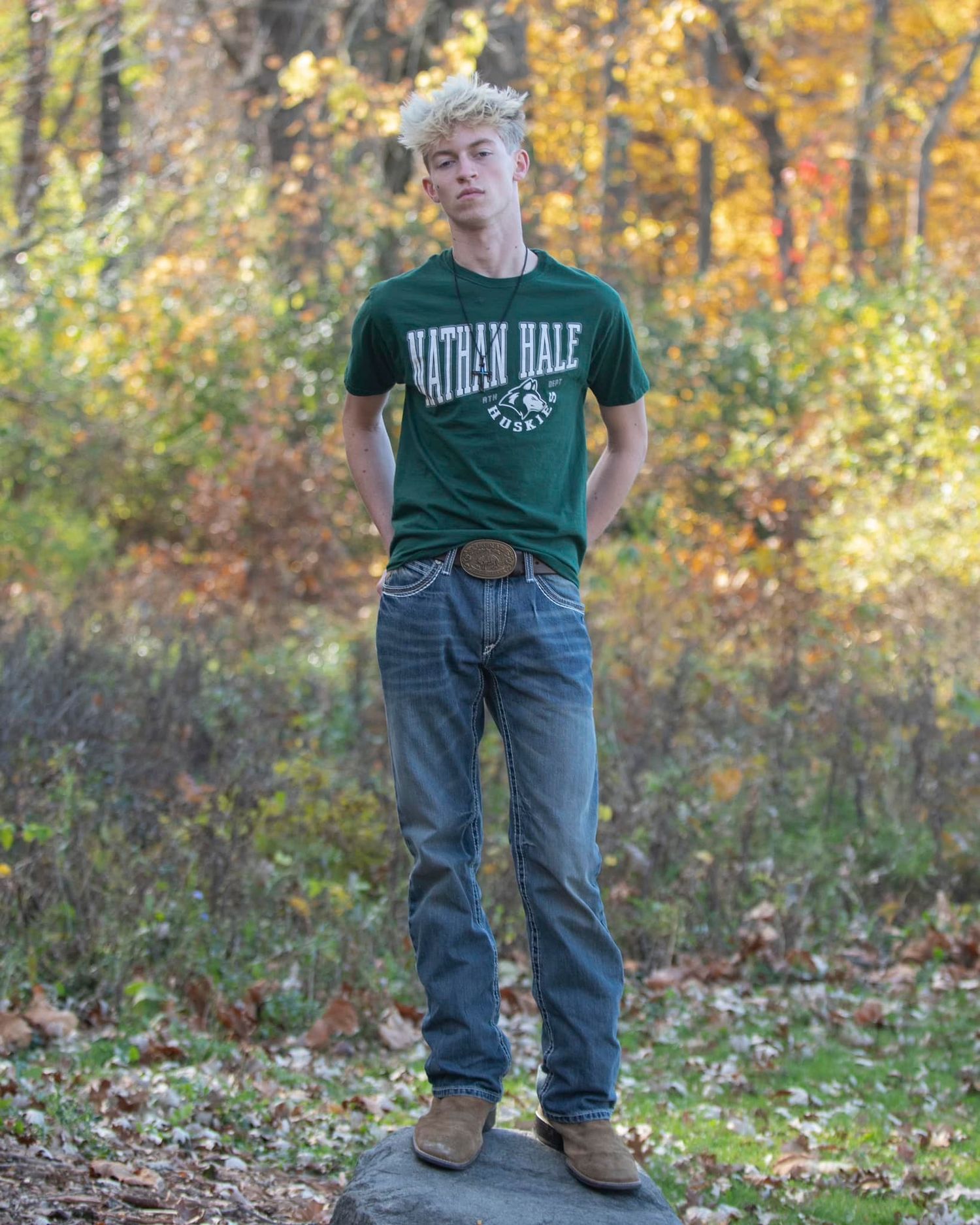 Young man in green Nathan Hale t-shirt and jeans standing on rocks surrounded by autumn foliage.