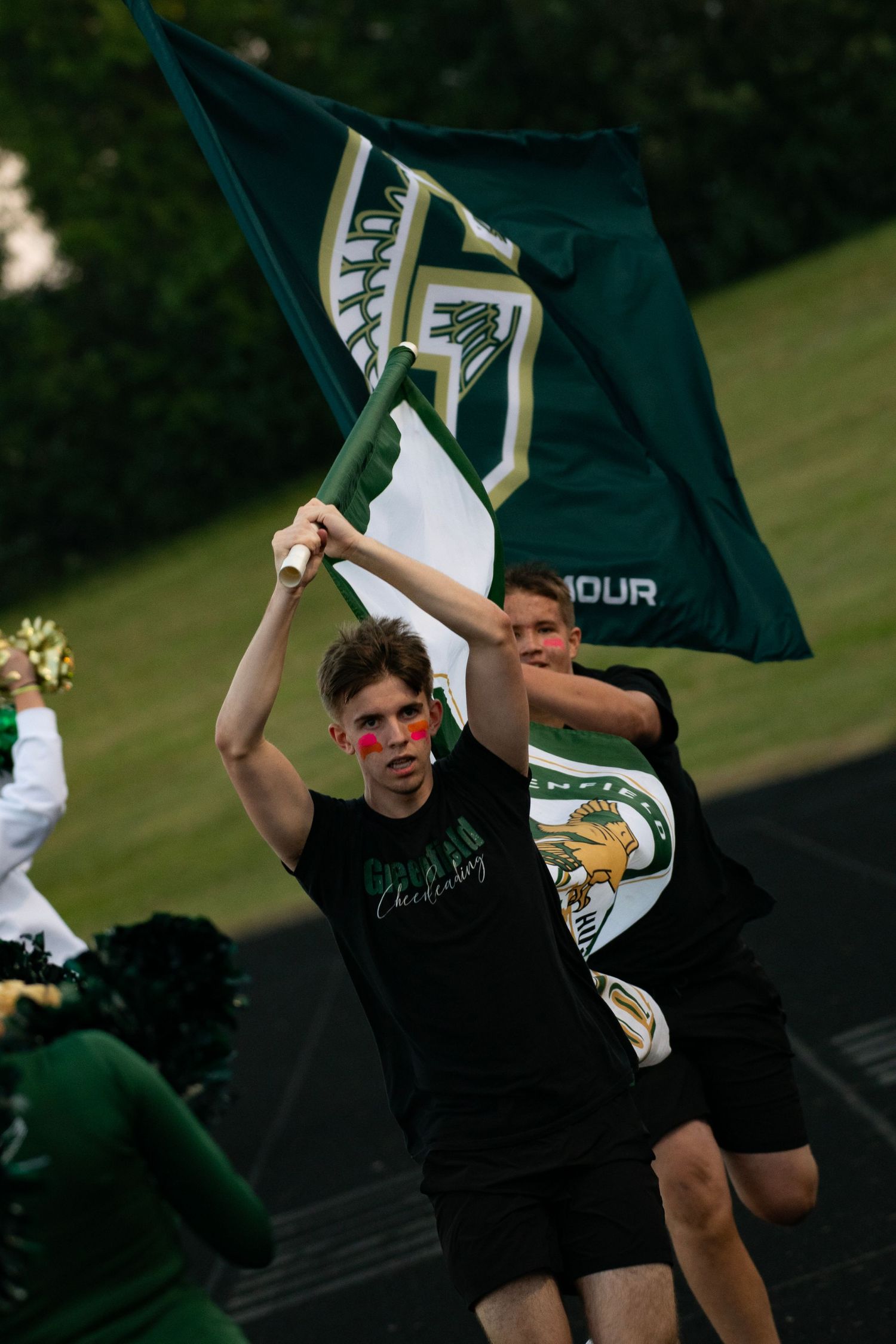 Young athlete runs with green and white school flag wearing face paint at a sporting event.