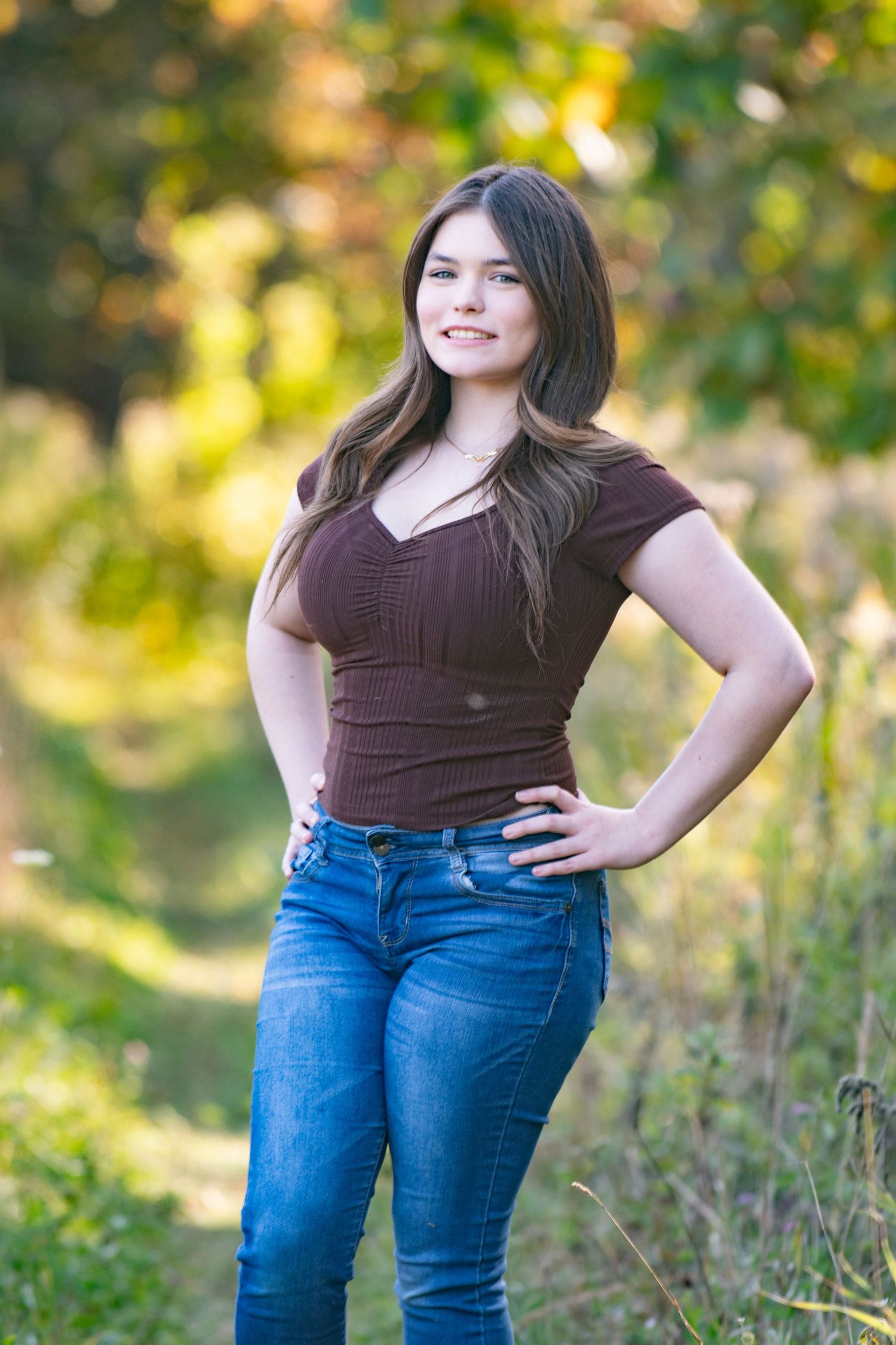 Someone wearing a brown shirt and blue jeans stands casually in front of lush greenery.