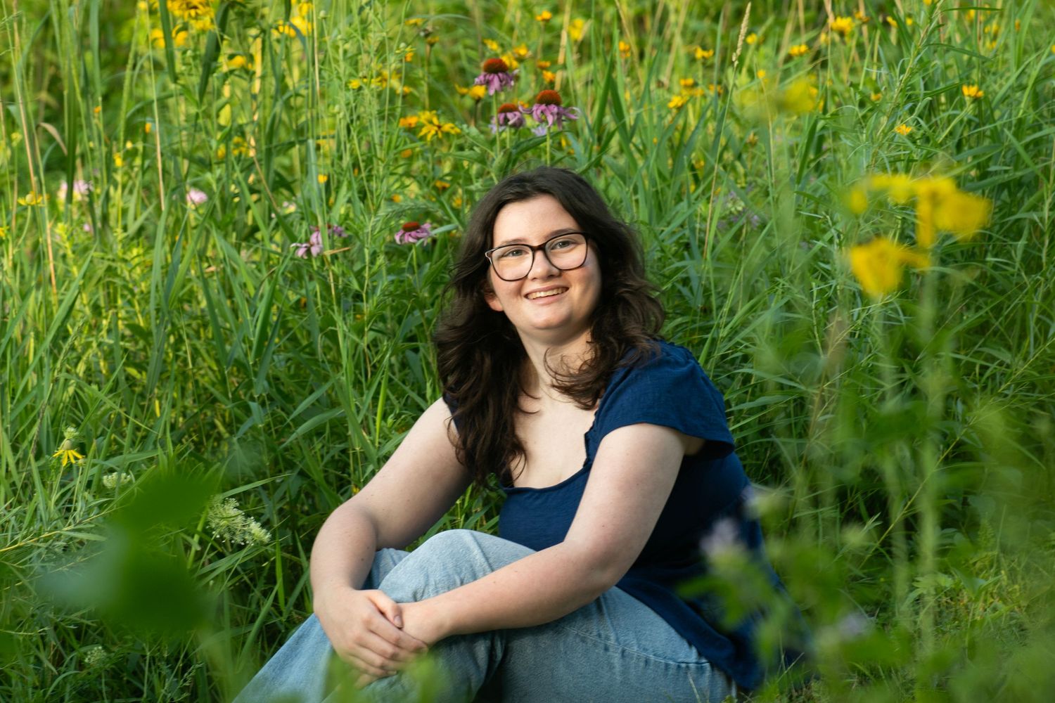 Young woman with glasses sitting in a lush green wildflower meadow, smiling warmly on a sunny day.