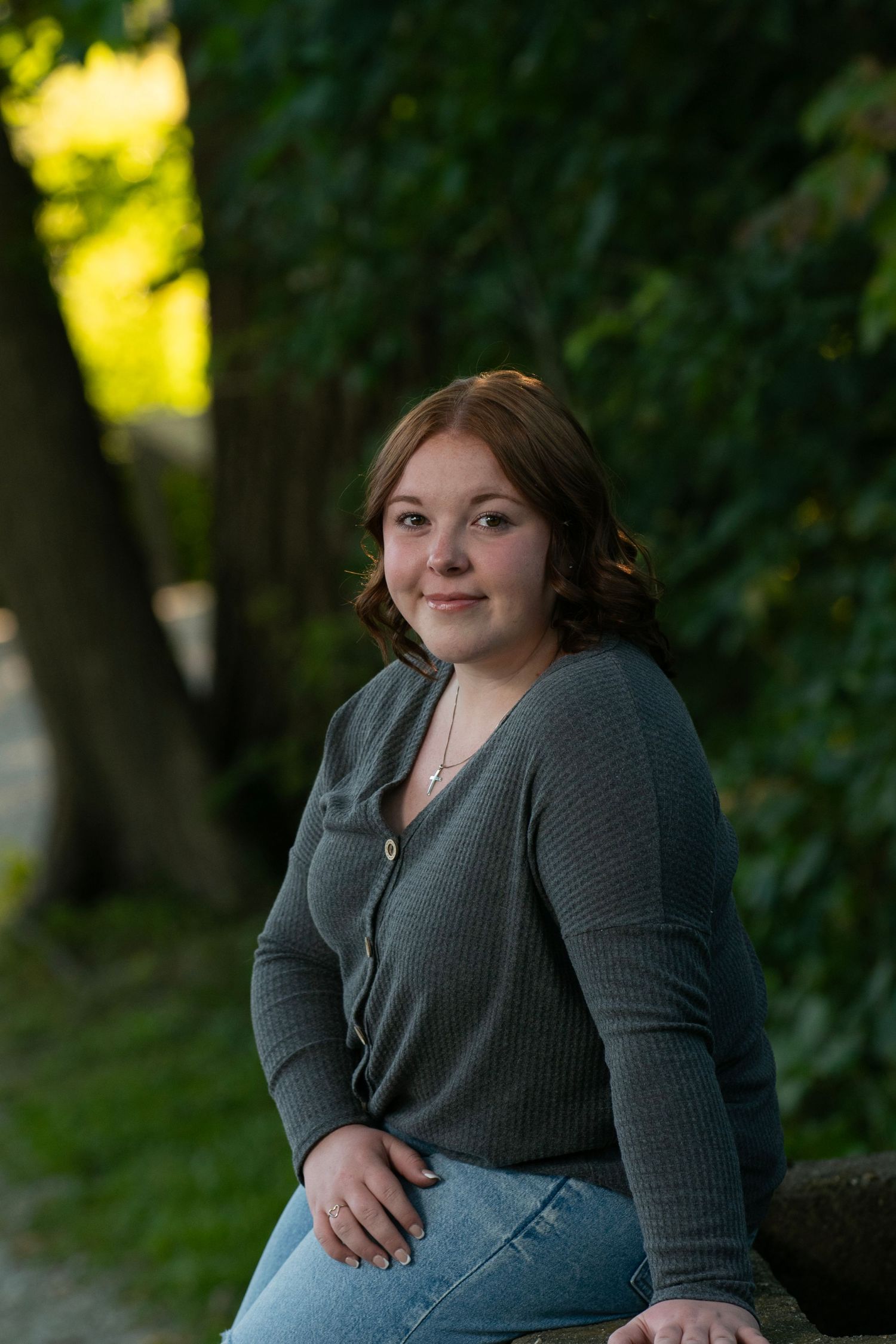 Young woman with red hair in gray ribbed top and jeans sitting outdoors among green trees smiling.