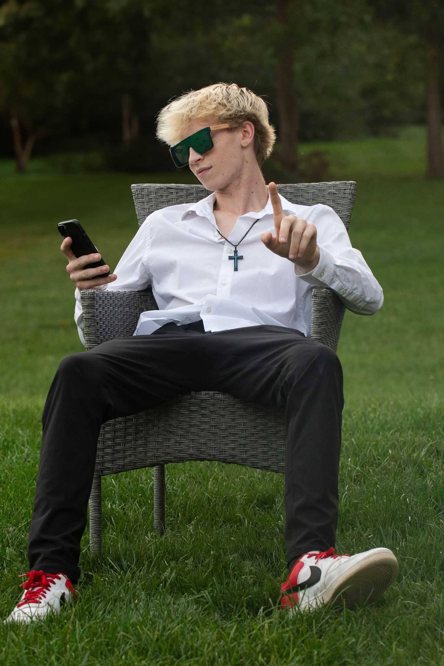 Young blond man in white shirt, green sunglasses, cross necklace sitting in wicker chair outdoors holding phone.