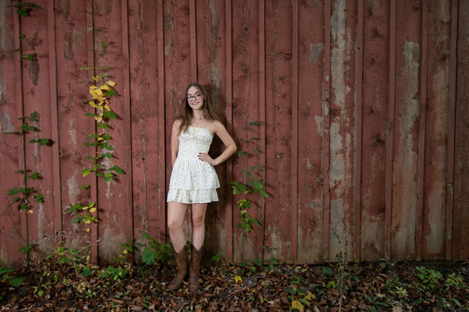 Young woman in white dress and boots posing against a rustic red barn wall with climbing vines.