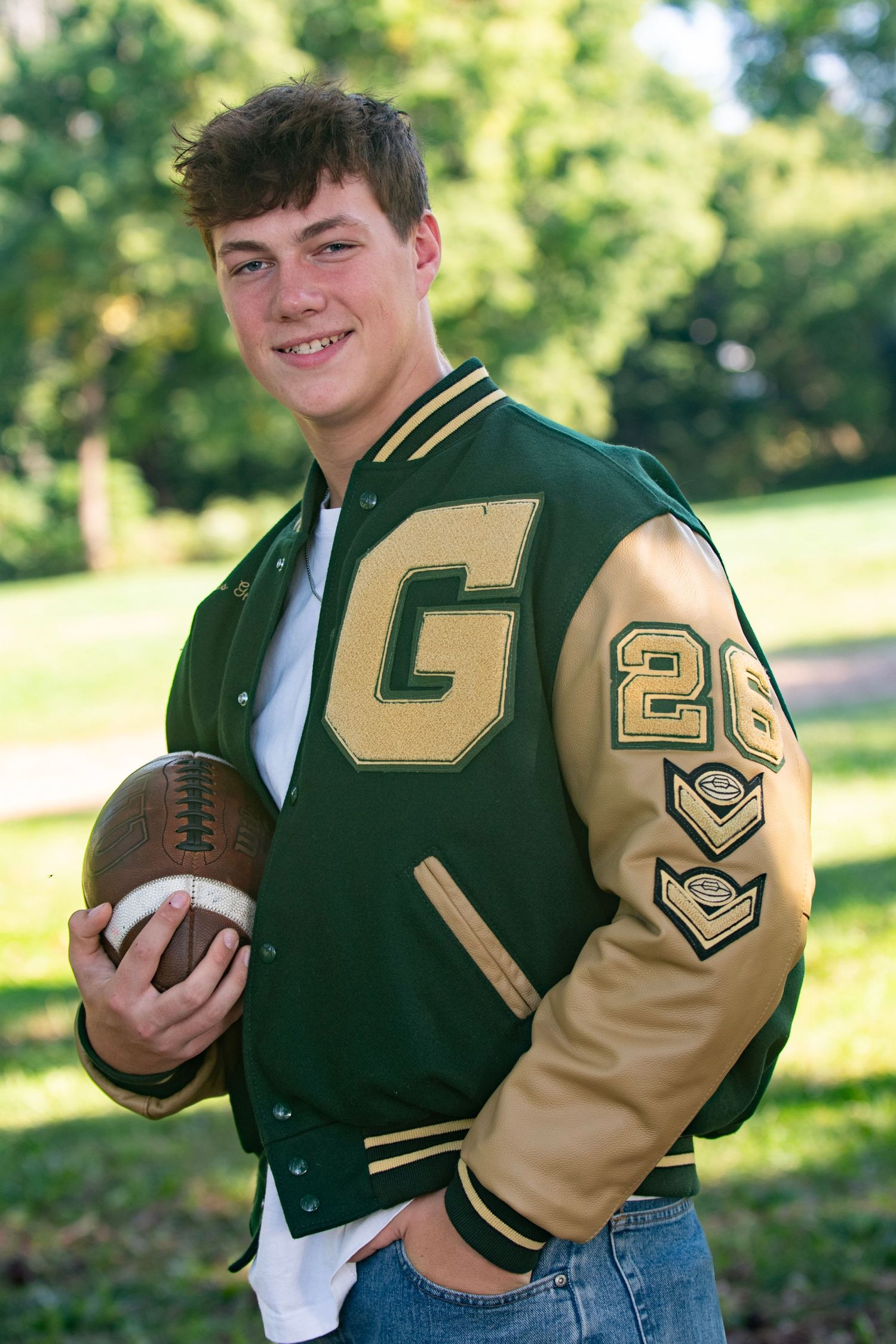 Young man in green and gold varsity jacket holding a football, wearing number 26, posing outdoors for a senior portrait.