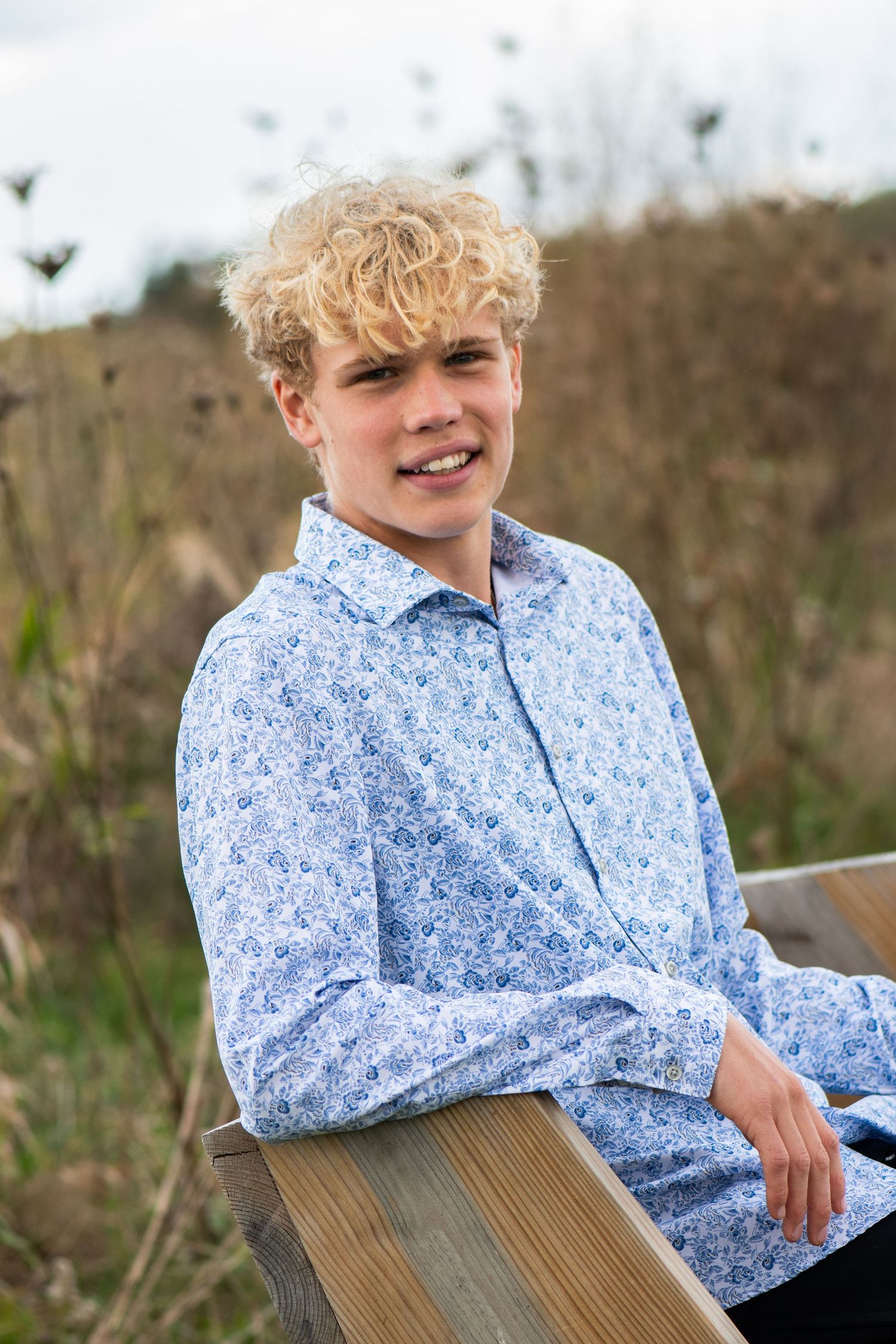 A casual outdoor portrait features someone in a blue floral shirt sitting on a wooden surface. Milwaukee County Grounds Game Day to Grad Day