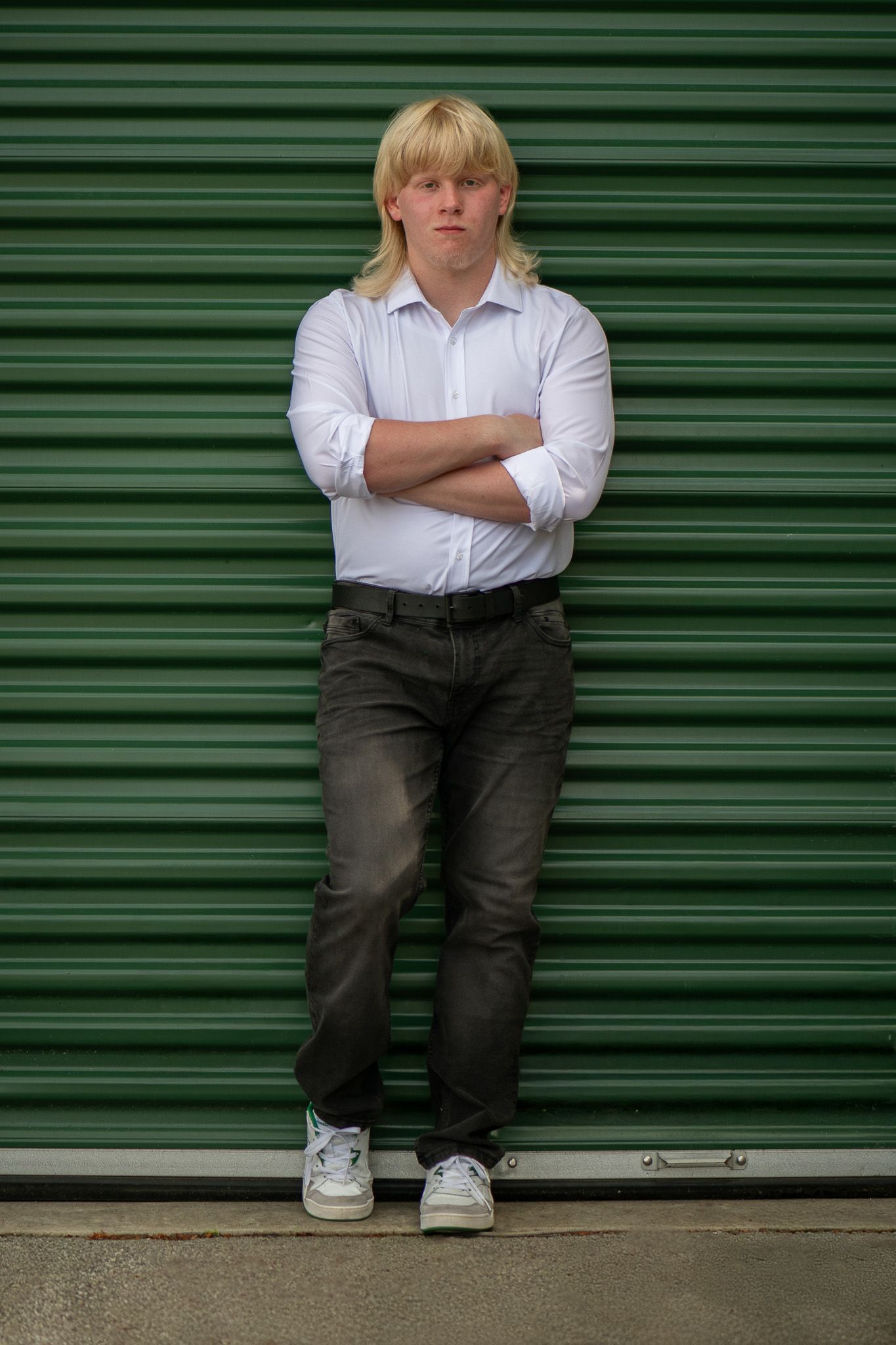 Person in white shirt and dark pants sitting against green metal storage door.