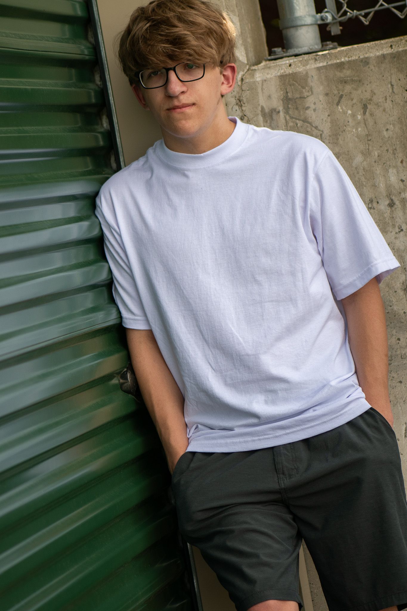 A casual portrait against a green storage door wearing glasses and white t-shirt for his senior portrait session in Greenfield, WI