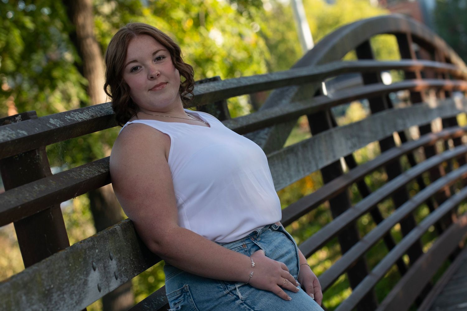 High school senior in a white top poses outdoors against a bridge in warm afternoon lighting for her Simply Junique Senior Portrait Session.