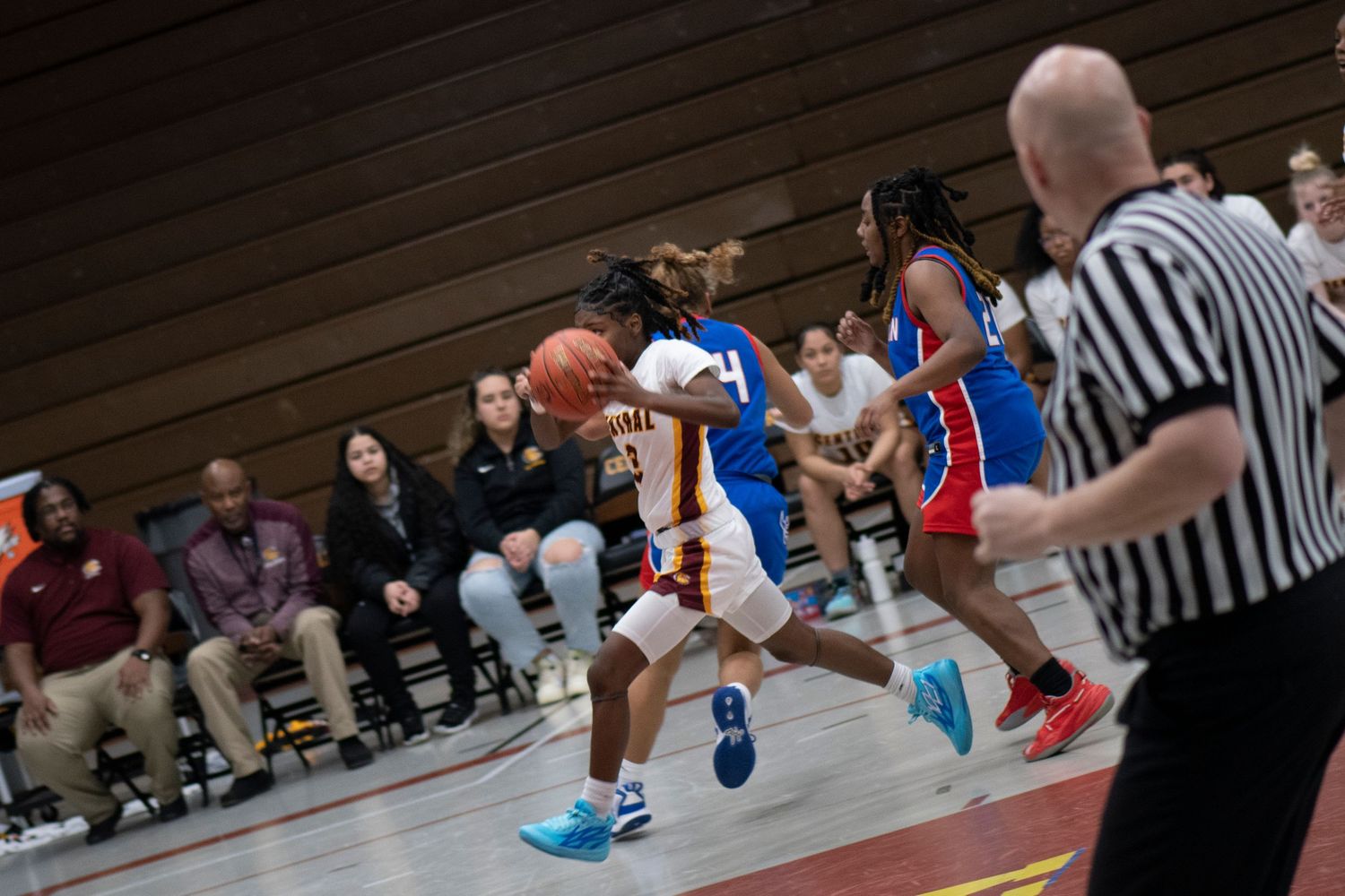 Basketball player dribbles down court during indoor game while referee and spectators watch from sideline.