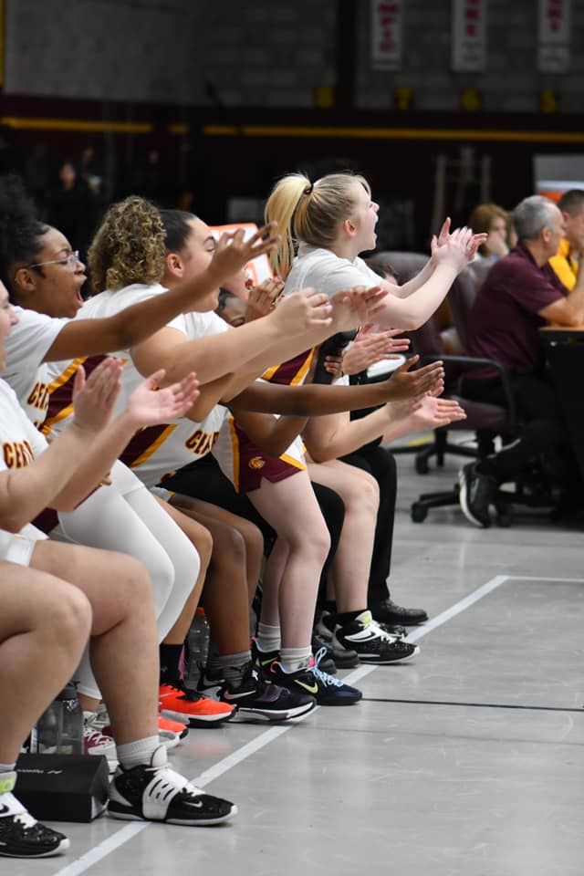 Women's basketball team bench players clapping and cheering in maroon and gold uniforms during a game.