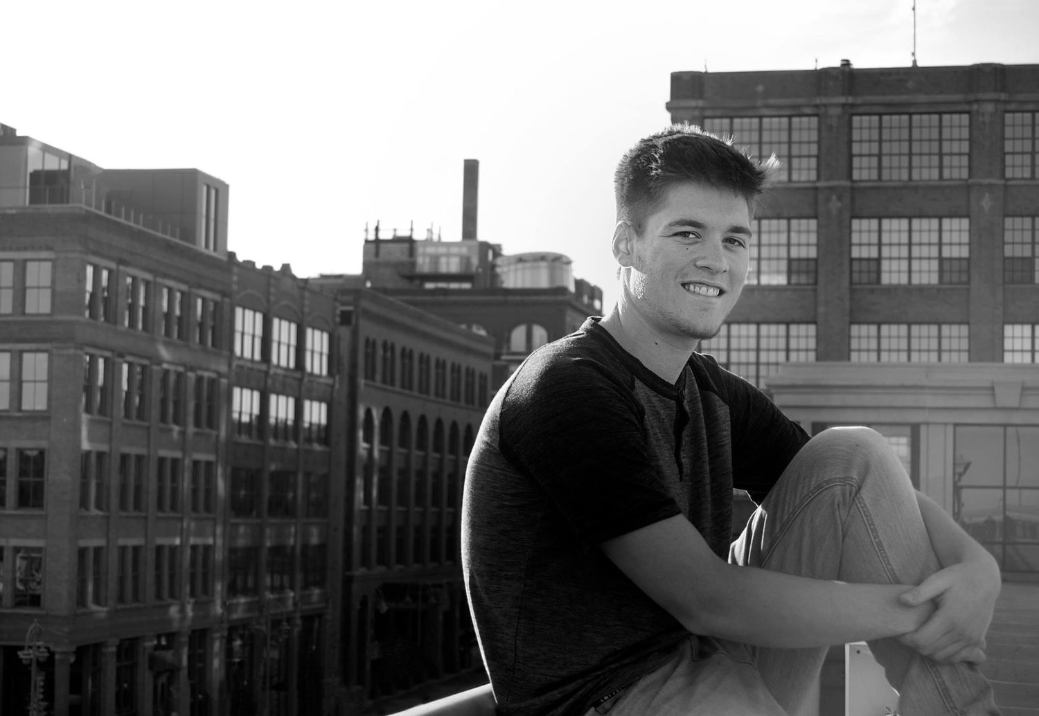 Young man smiling on rooftop with urban brick buildings in black and white photo.