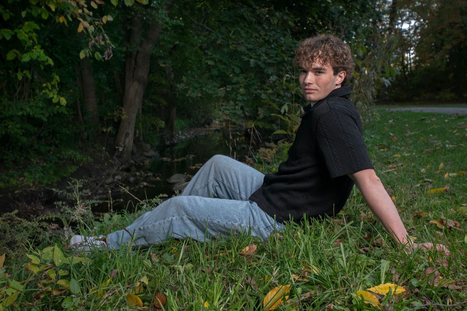Young man in black shirt and jeans sitting on grass near trees, looking back over his shoulder outdoors.