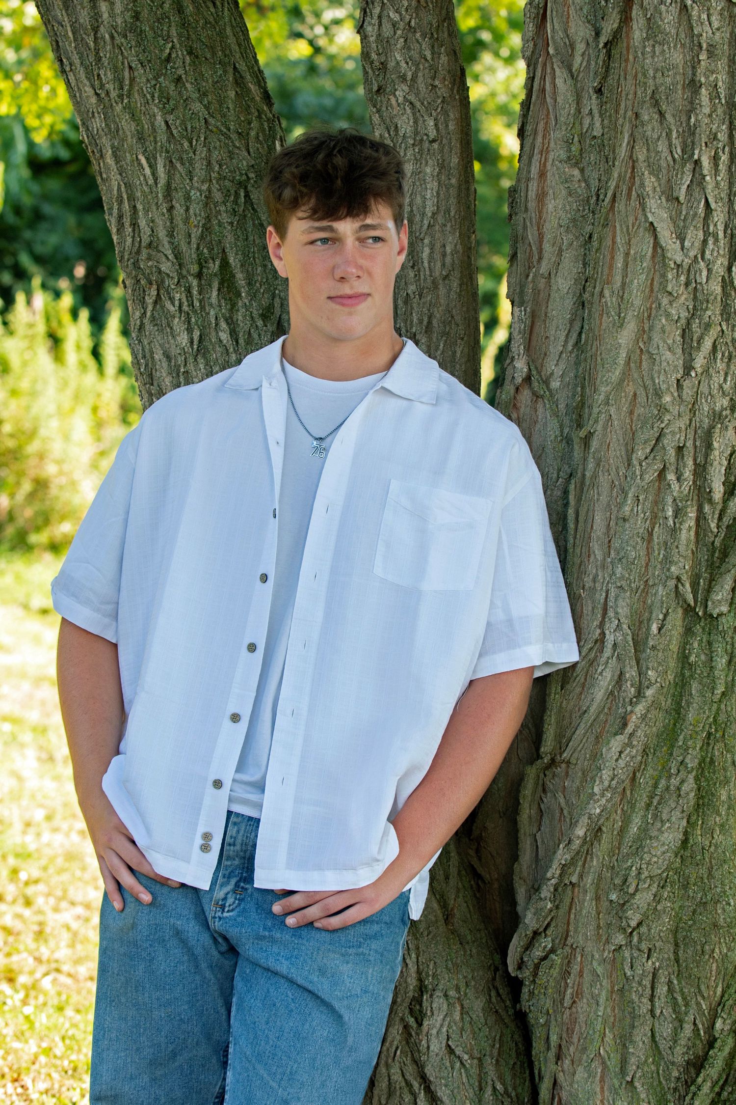 A person in a white button-up shirt and jeans poses casually against a tree trunk in a sunlit outdoor setting.