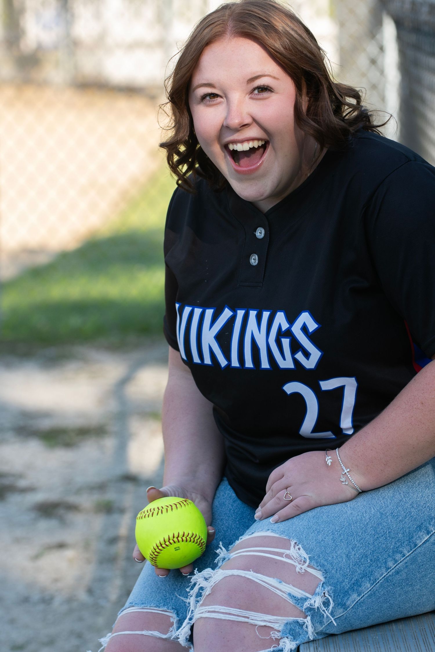 High school athlete wearing a Vikings jersey sits outdoors with a softball, displaying ripped jeans and her fun personality.
