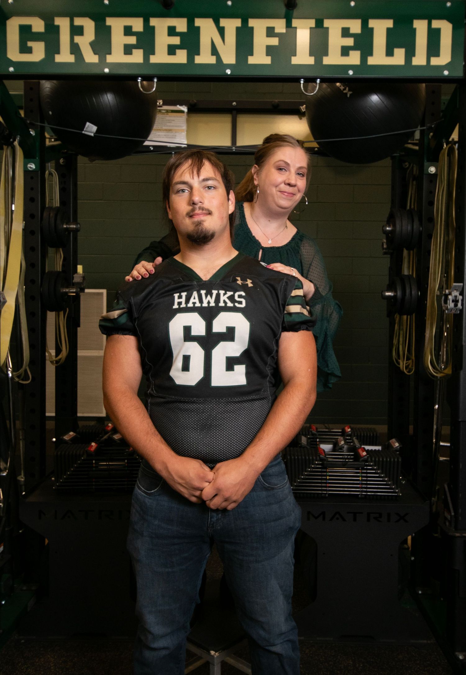 Four athletes in black jerseys and jeans stand together in a weight room with workout equipment visible in the background.