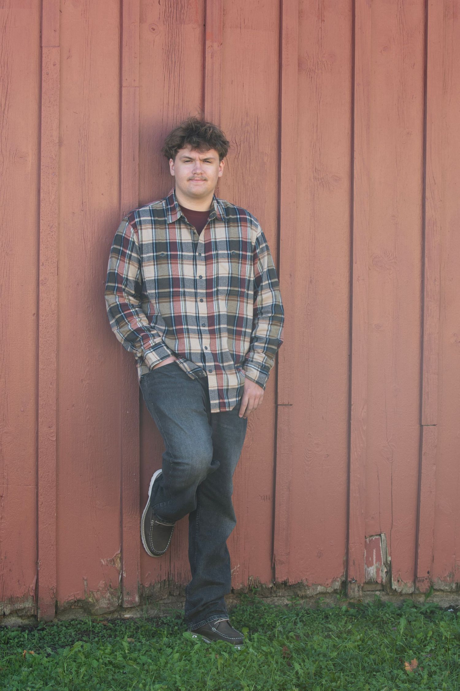 Young man in plaid flannel shirt and jeans leaning against a rustic red barn wall on green grass.