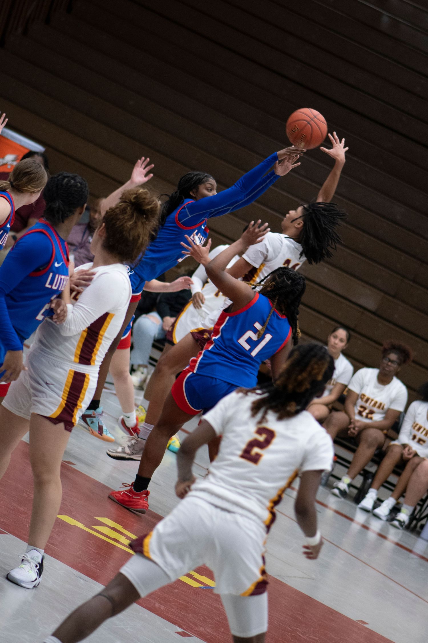 Women's basketball game action shot with players in blue and white uniforms competing for a rebound near the basket.