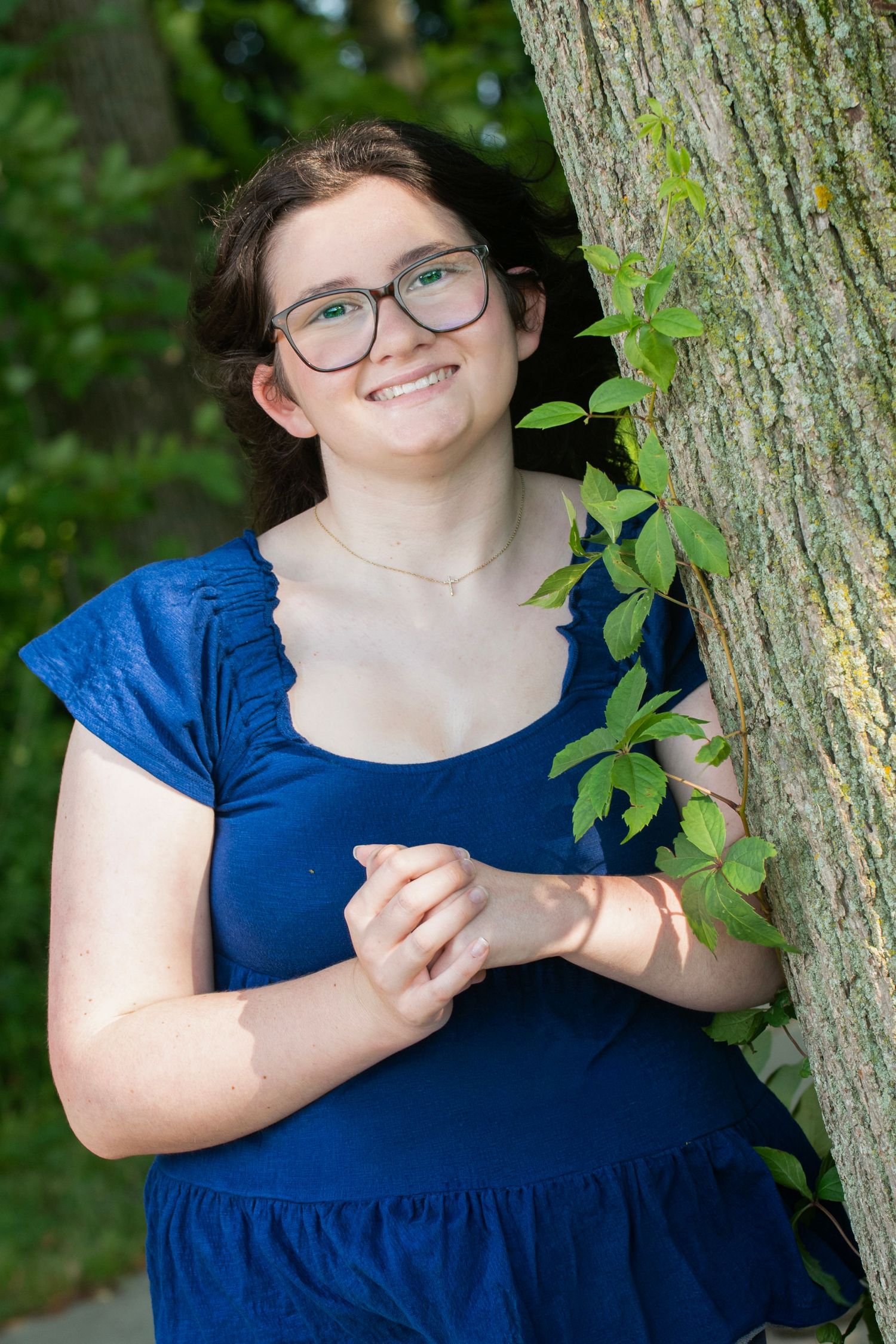 Smiling young woman with glasses in blue dress leaning against a tree outdoors.