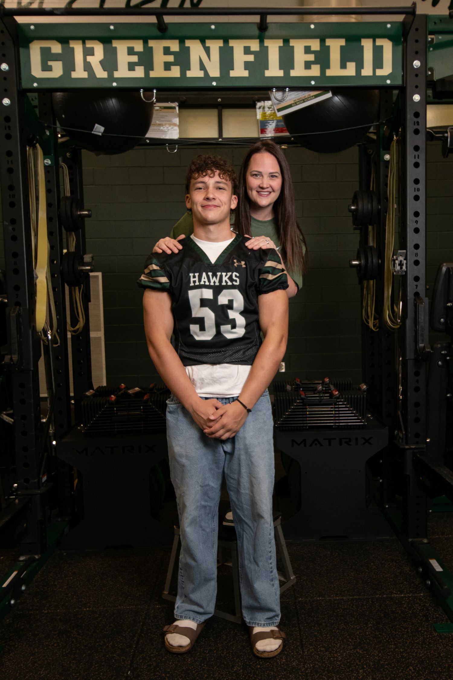 Two people wearing Hawks jerseys stand together in a dimly lit weight room with Greenfield signage visible.