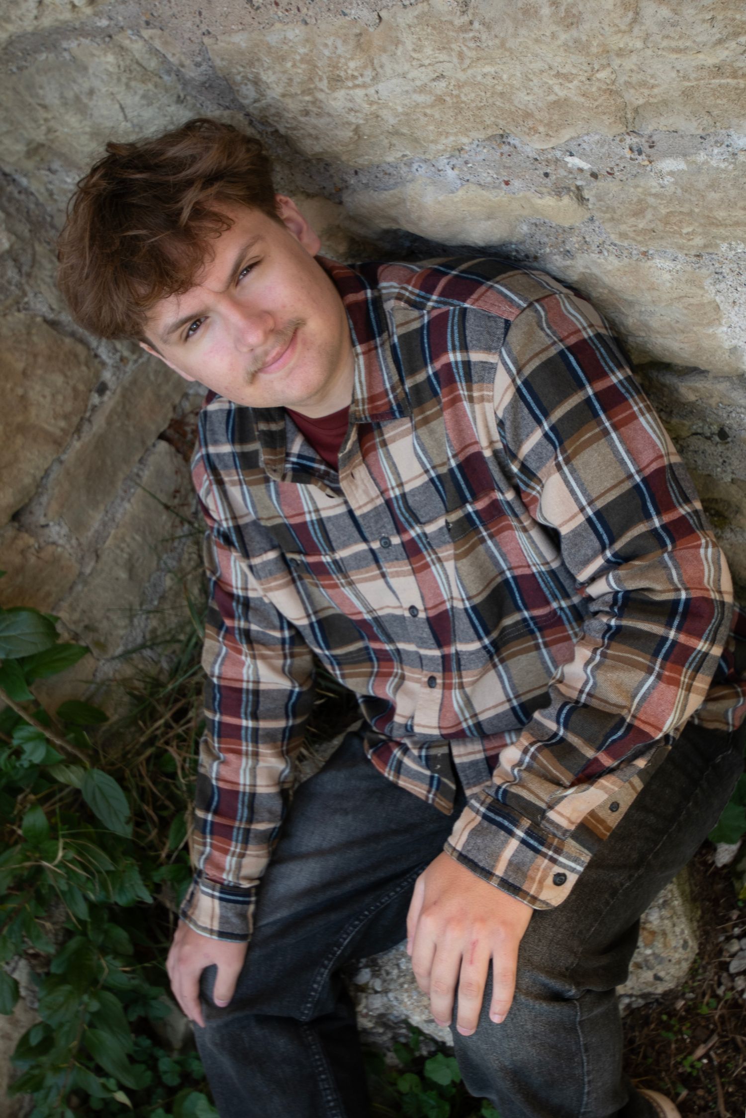 Young man in plaid flannel shirt leaning against stone wall outdoors.