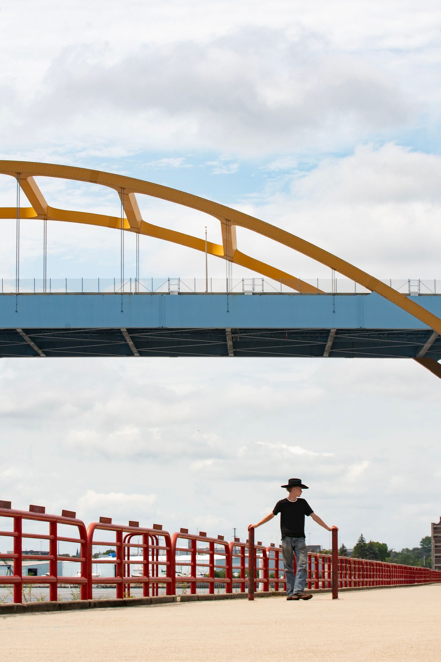 Person in black hat stands on red-railed bridge walkway beneath yellow arch bridge against cloudy sky.