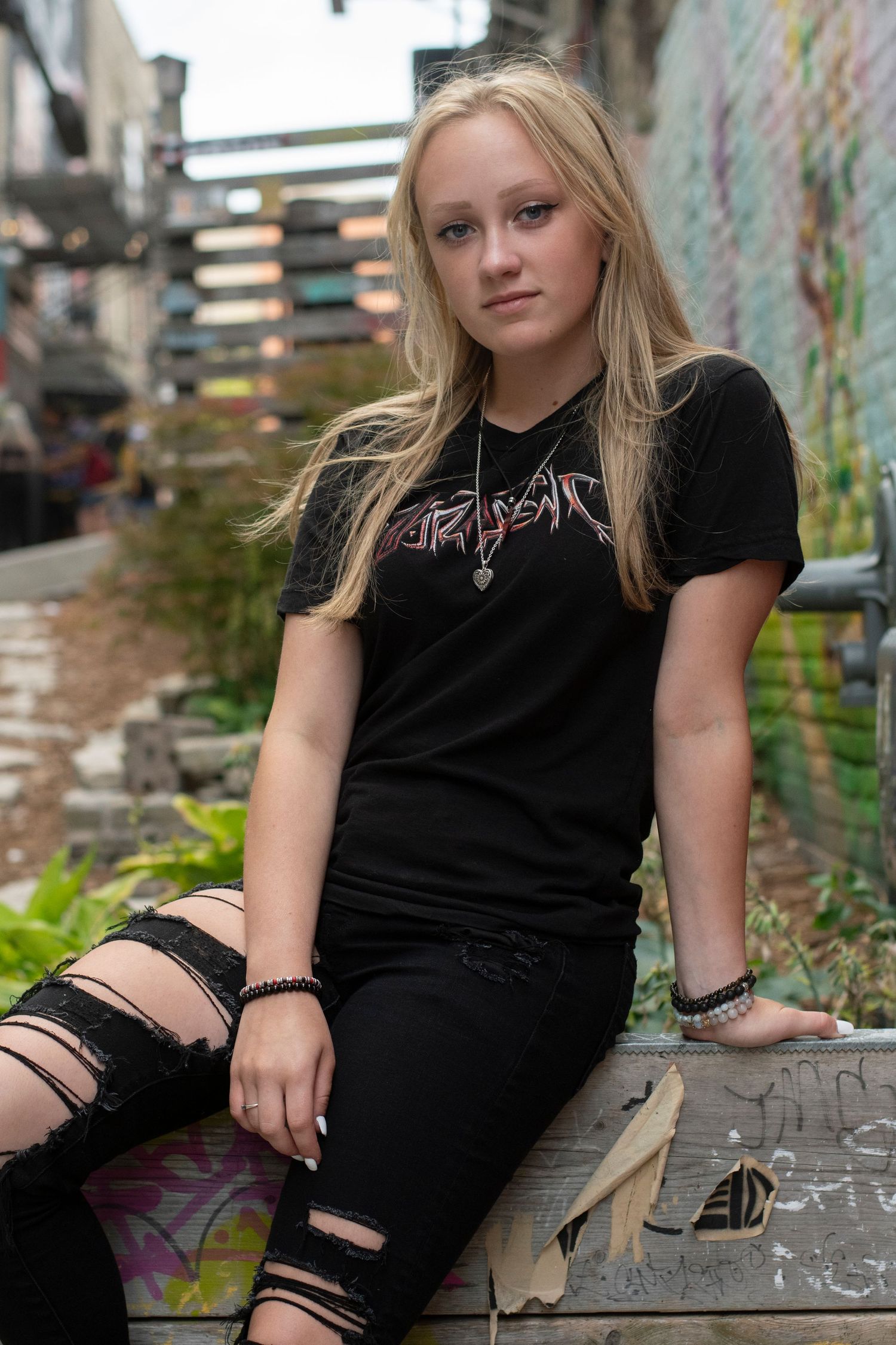 Young blonde woman in black ripped jeans and band tee sits in urban alley with graffiti walls.