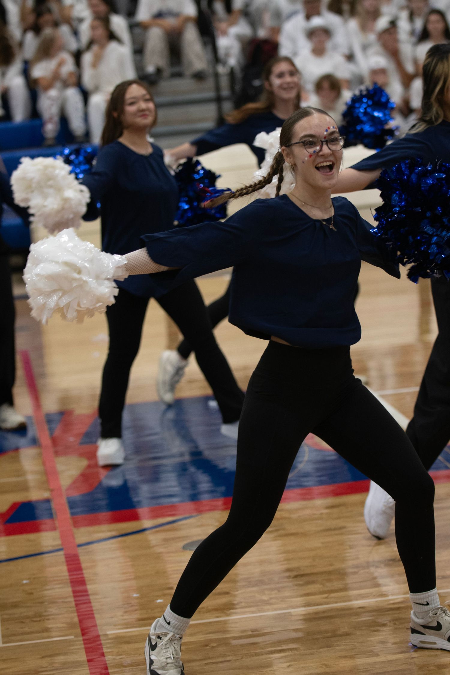 Dance team members in black outfits perform an energetic routine on a gymnasium floor with blue and white pom poms.