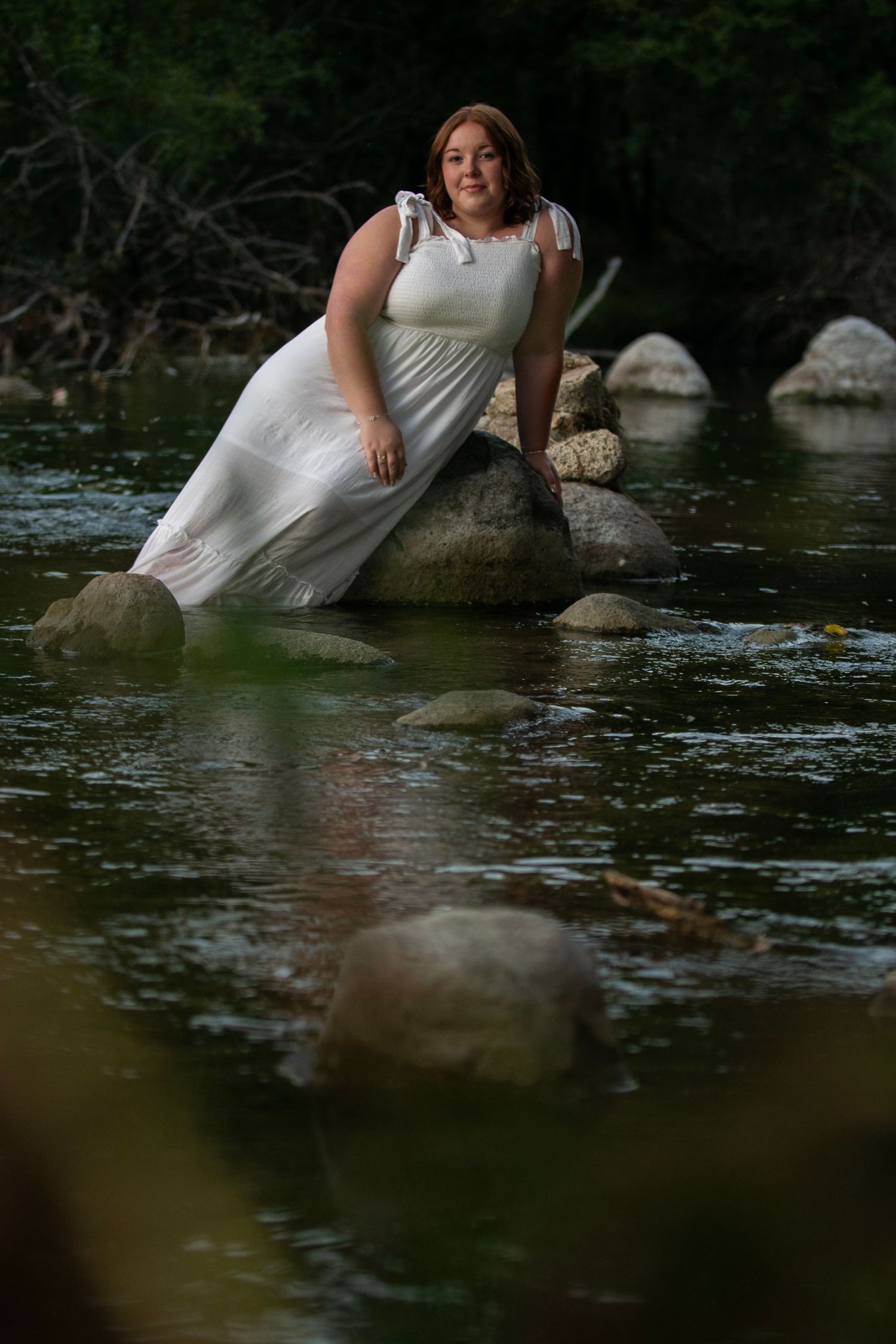 A high school senior in a white dress poses on rocks in a flowing river during a nature photoshoot during Simply Junique senior session.