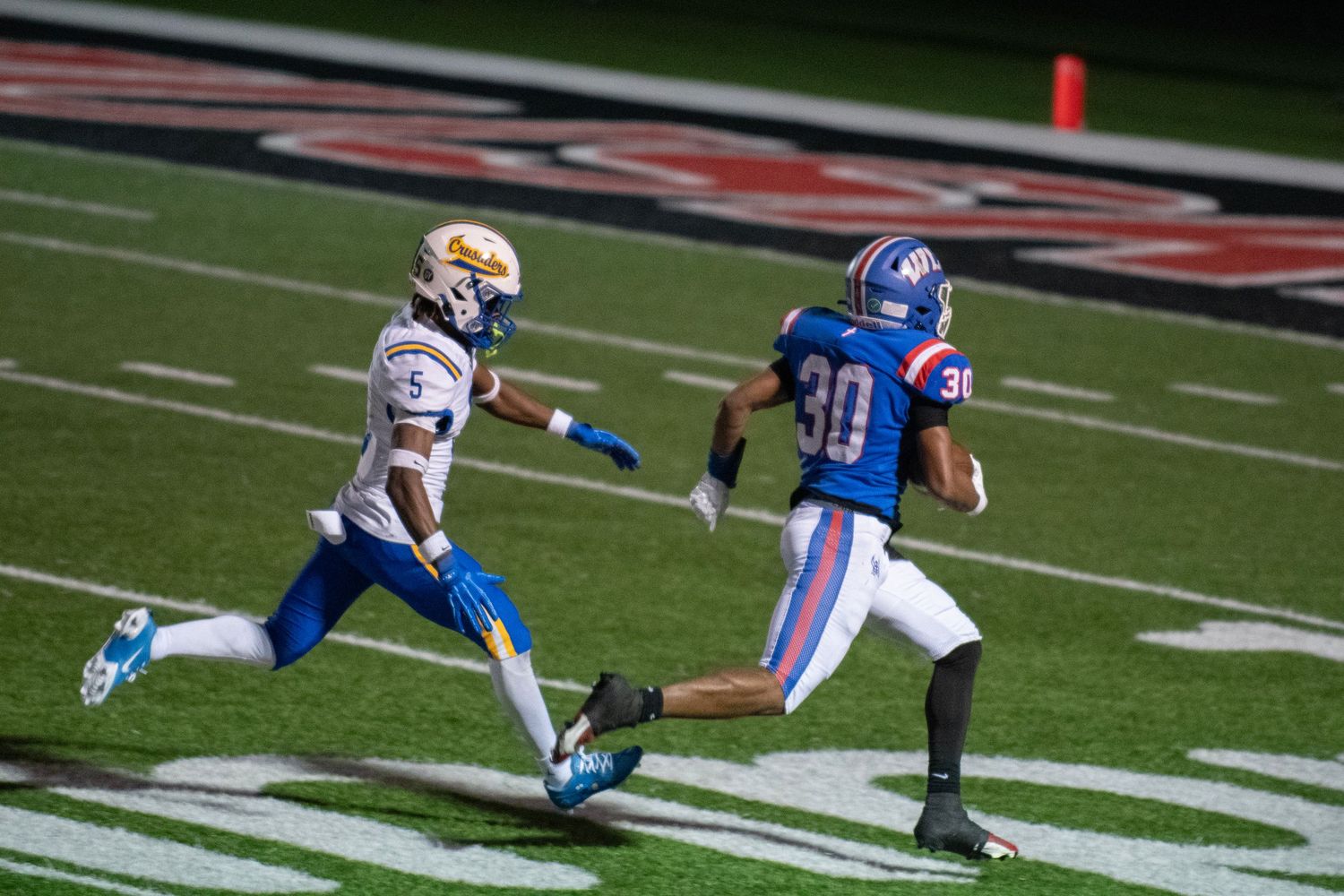 Two football players race down the field at night, one in blue number 30 jersey, the other in white and gold uniform.