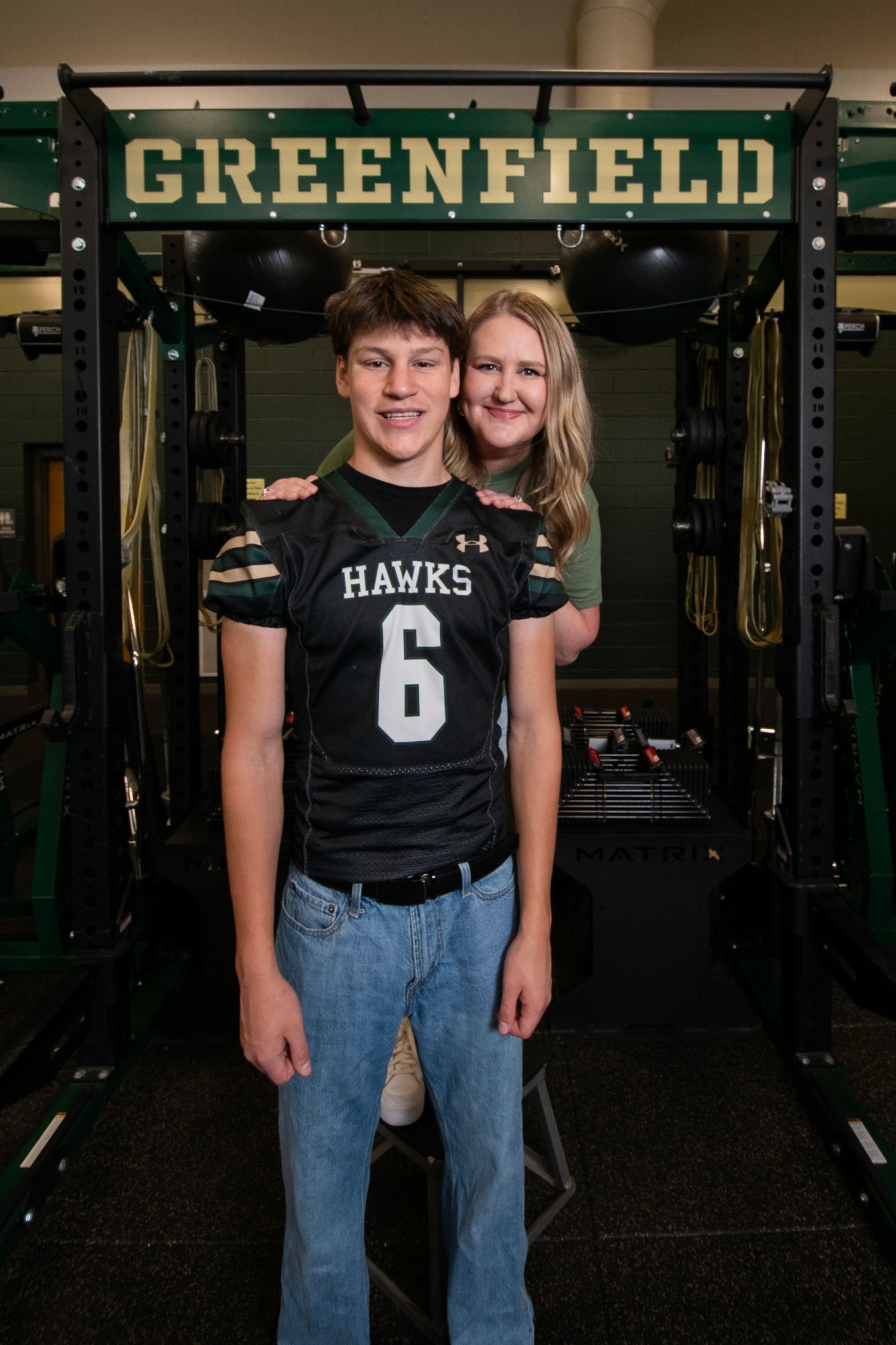 Student athletes pose in front of Greenfield weight room wearing Hawks jersey number 6 and jeans.