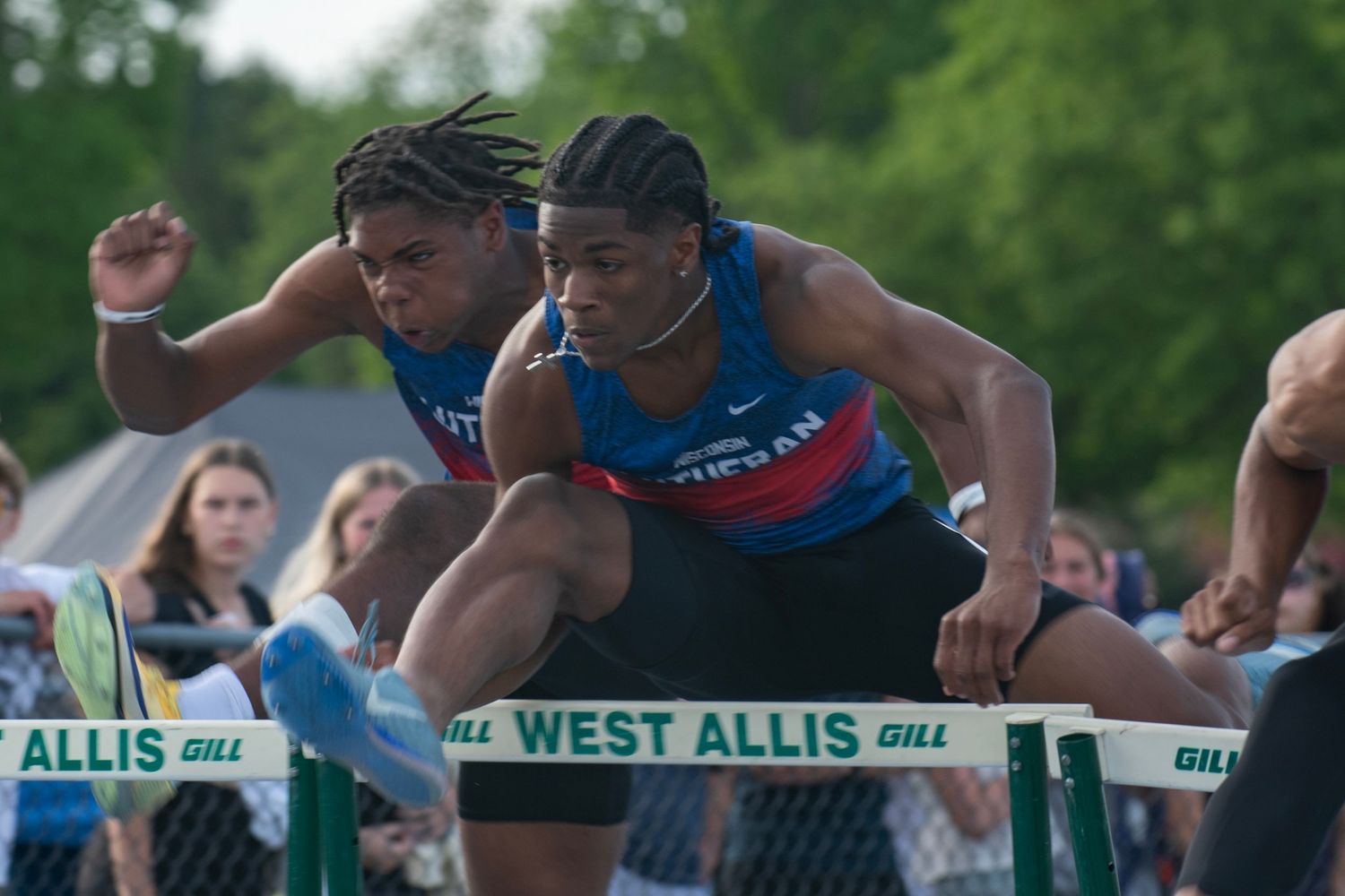 Two athletes compete in a hurdles race at a West Allis track meet, leaping over barriers in athletic competition.