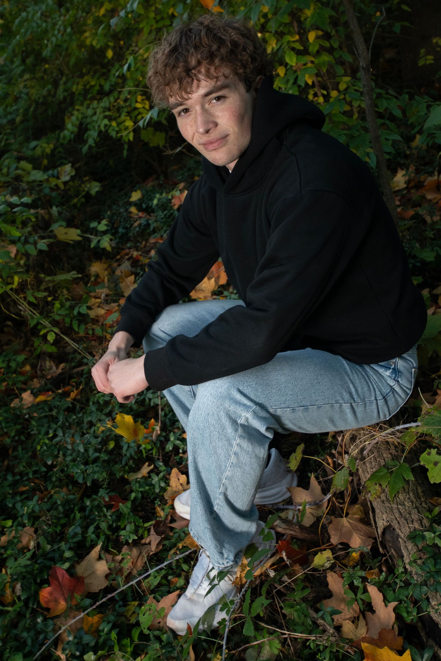 Teen boy in black hoodie and jeans sits on log surrounded by green ivy and fall leaves.
