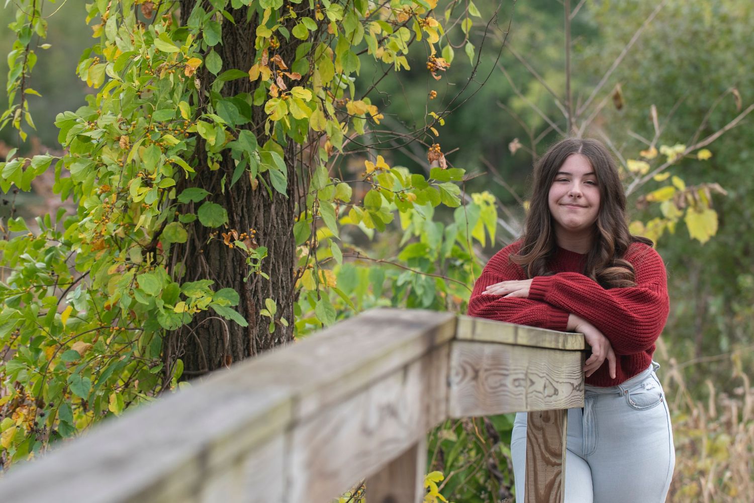 A smiling person in a cozy red sweater and jeans poses against autumn foliage and a rustic wooden fence.