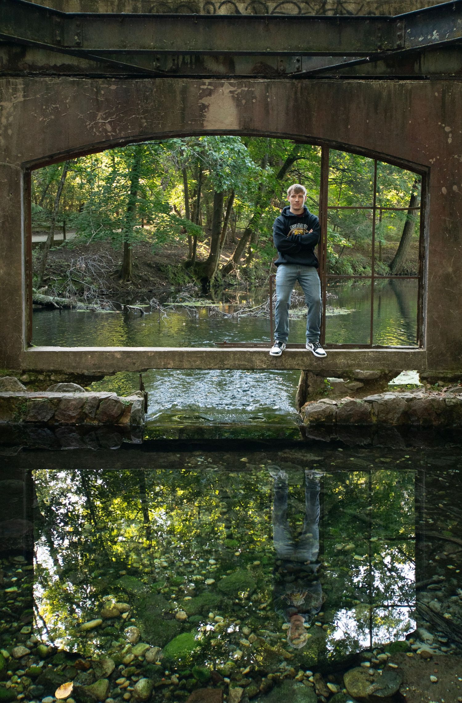 A figure stands framed by a stone bridge archway over a tranquil stream with trees and reflections in the water below.