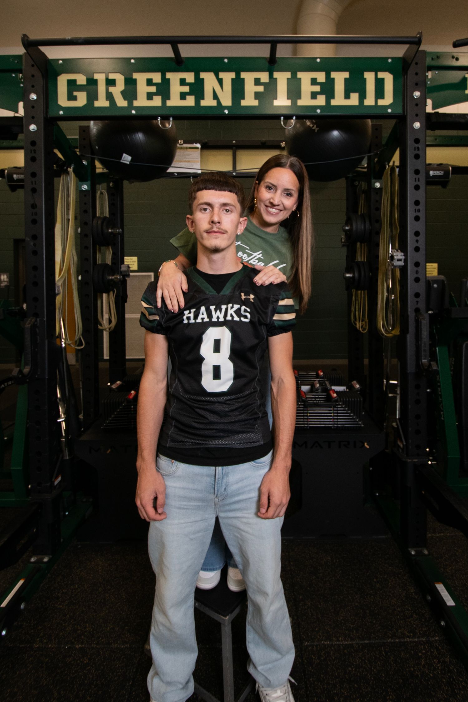 Athletes in black Hawks jerseys and light jeans pose in a green and black weight room with workout equipment.