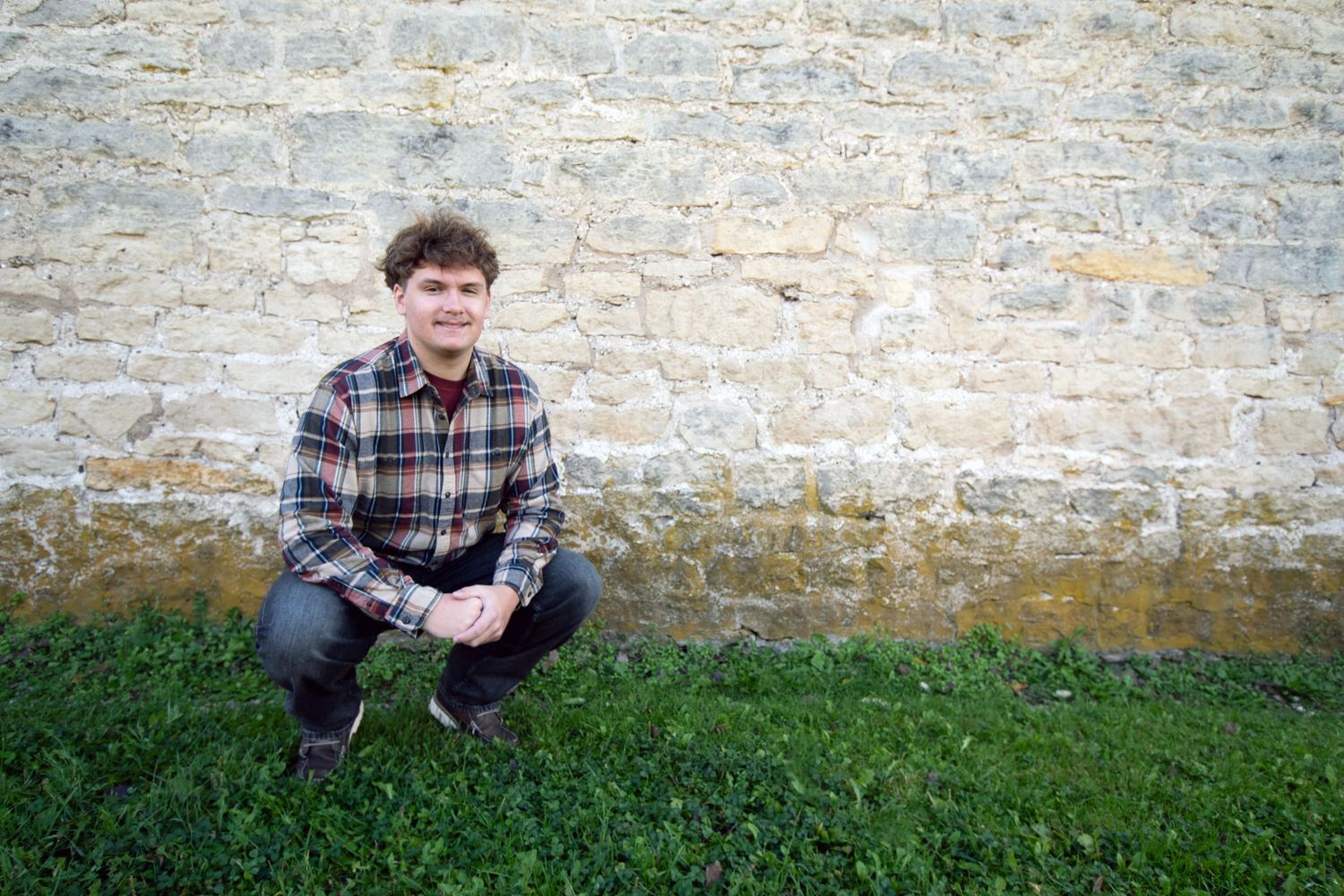 Young man in plaid shirt crouching on green grass against a rustic stone wall outdoors.