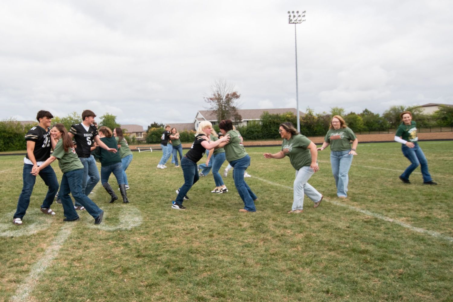 People running and playing together in a line across a grassy sports field under cloudy skies.