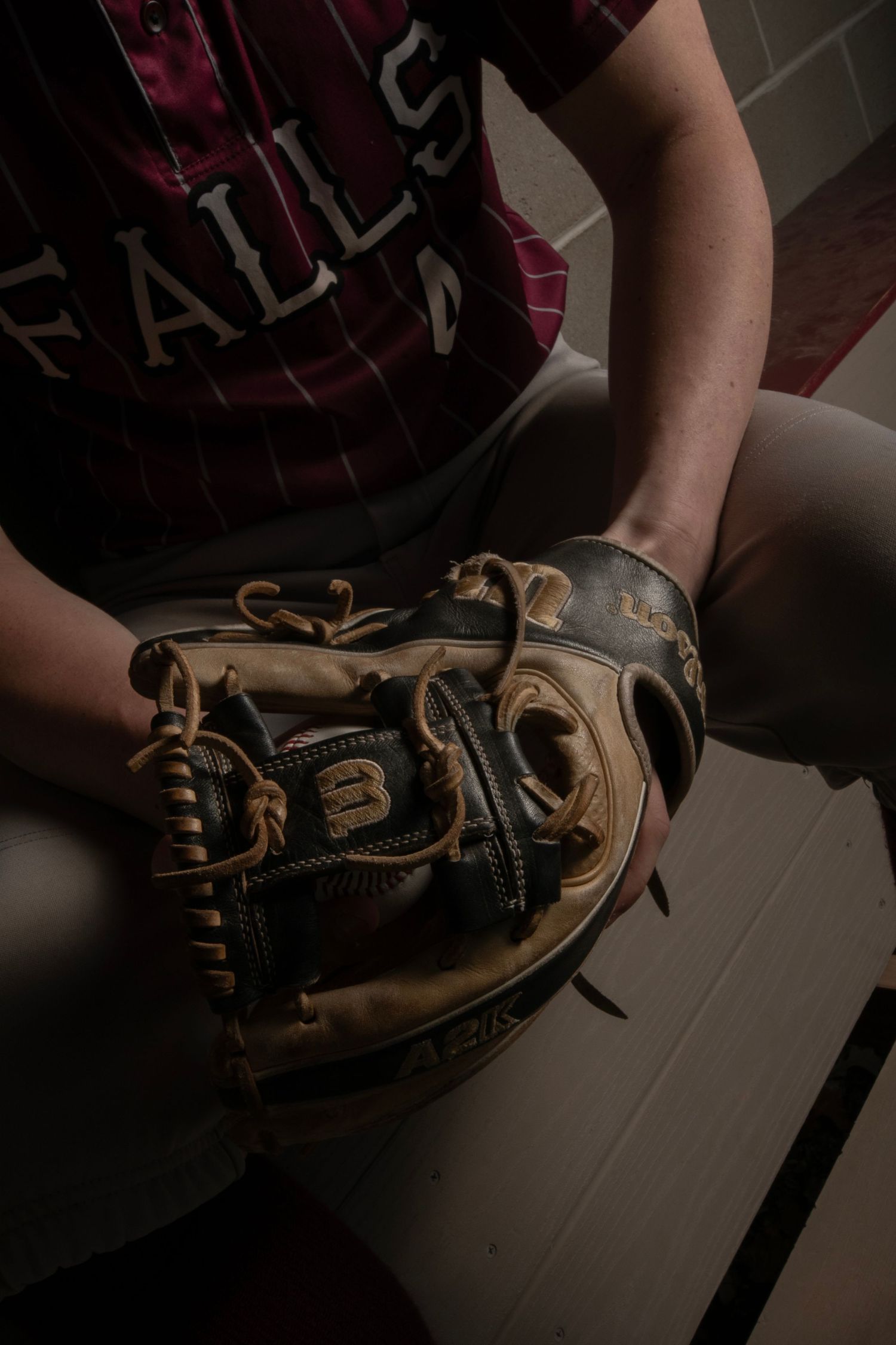 Baseball player in a Falls jersey holding a worn Wilson fielding glove while sitting in a dugout.