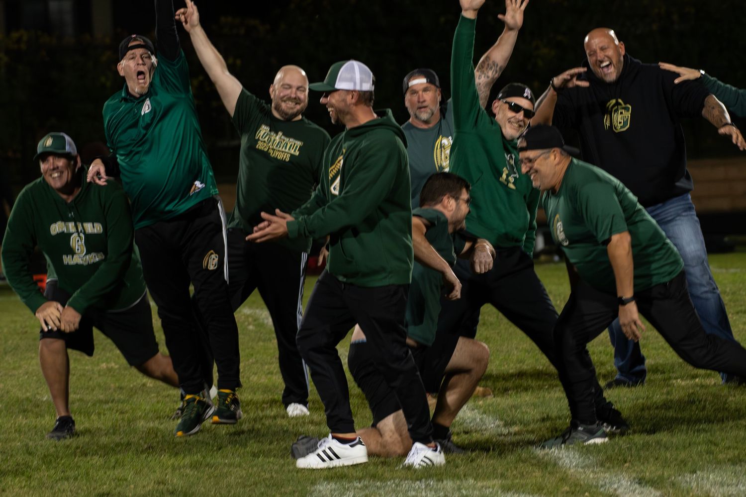 A group of people in green sports attire pose playfully together on a field at night.