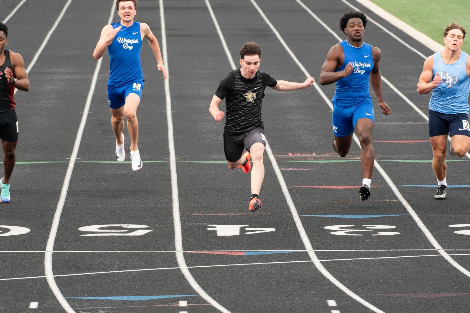 Competitive sprinters racing on a track, with a runner in black leading the pack during a high school track and field meet.