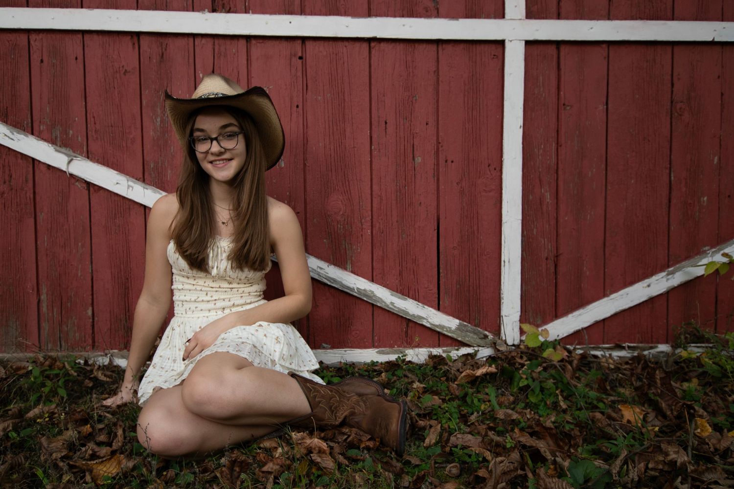 A rustic photo session against a red barn wall with white trim features someone in a white lace dress and cowboy hat.