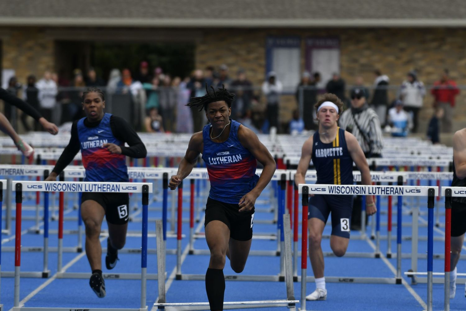 Athletes compete in a hurdles race on a blue track at a Wisconsin Lutheran track and field meet.